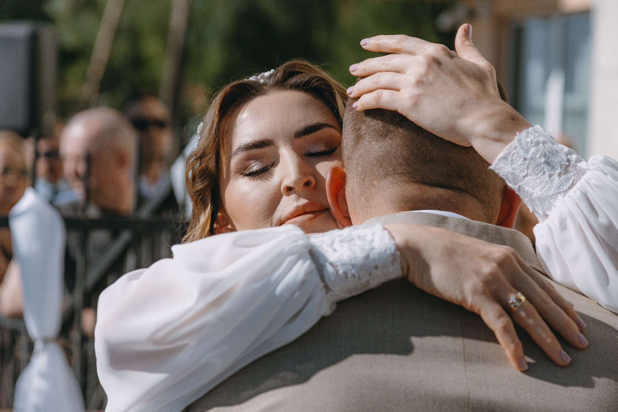 Ujęcie panny młodej z zamkniętymi oczami, obejmującej pana młodego. Delikatny makijaż, perłowa ozdoba we włosach, koronkowy mankiet. W tle rozmyte sylwetki gości.