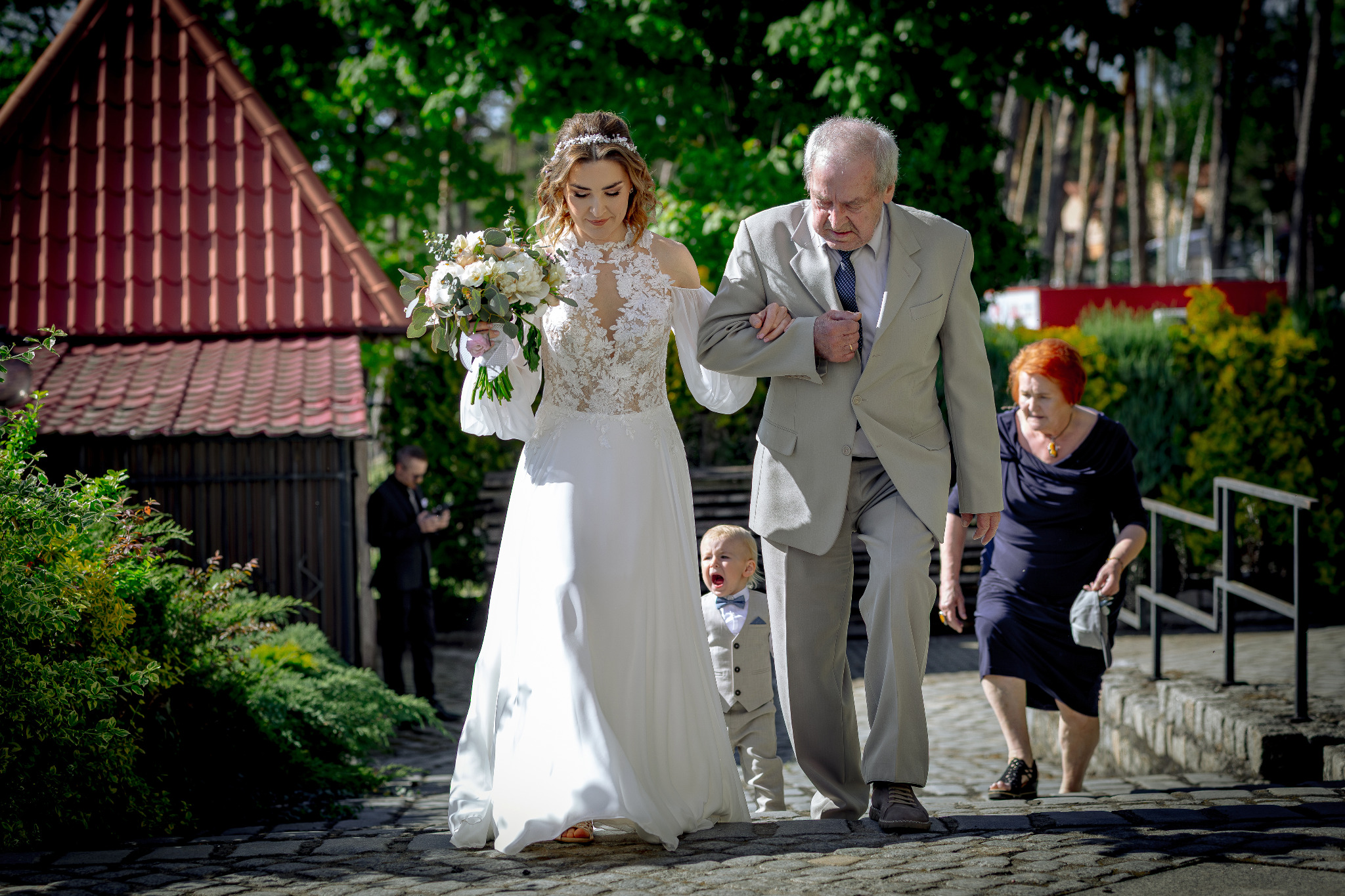 Panna młoda w koronkowej sukni prowadzona do ołtarza przez starszego mężczyznę, z płaczącym chłopcem w garniturze i gośćmi w tle, na tle zieleni i architektury.
