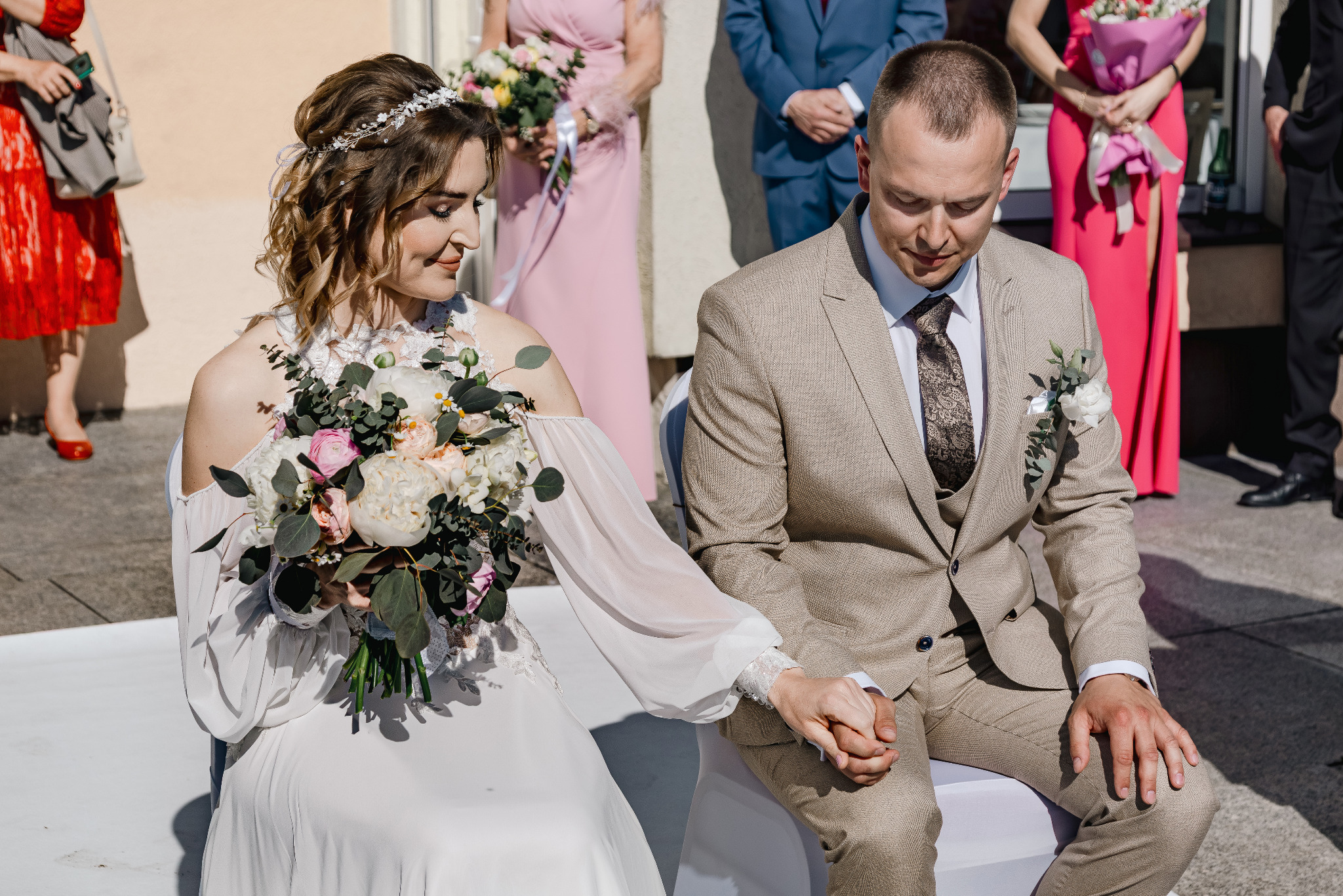 Para młoda siedzi obok siebie, trzymając się za ręce podczas ceremonii ślubnej. W tle drużba i druhny w pastelowych strojach.