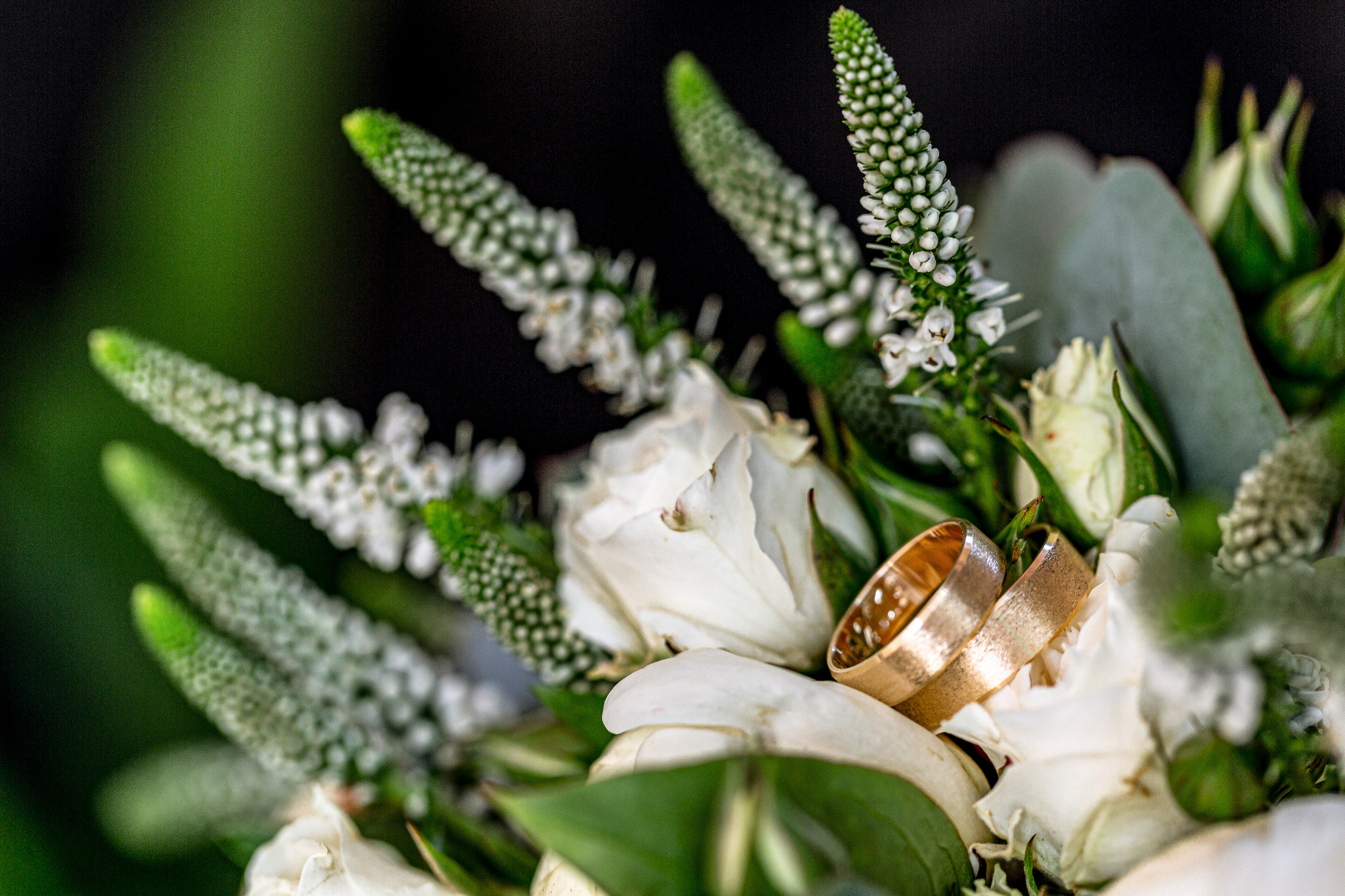 Złote obrączki ślubne spoczywają na bukiecie z białych róż i zielonych dodatków, tworząc romantyczną kompozycję. Makro, delikatne światło.