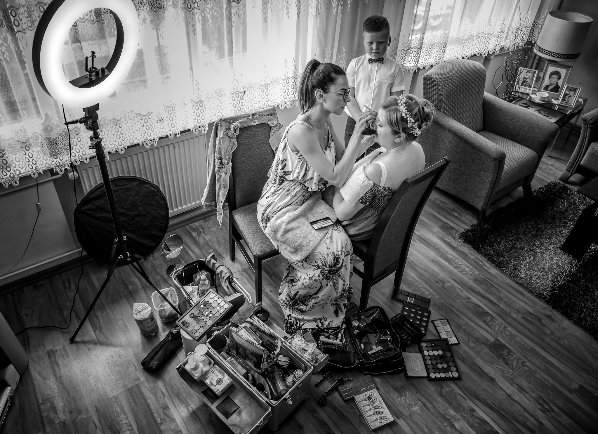 Wizażystka w trakcie pracy nad makijażem panny młodej, w tle asystujący chłopiec w muszce. Widoczny sprzęt oświetleniowy i kosmetyki. Czarno-biała fotografia.