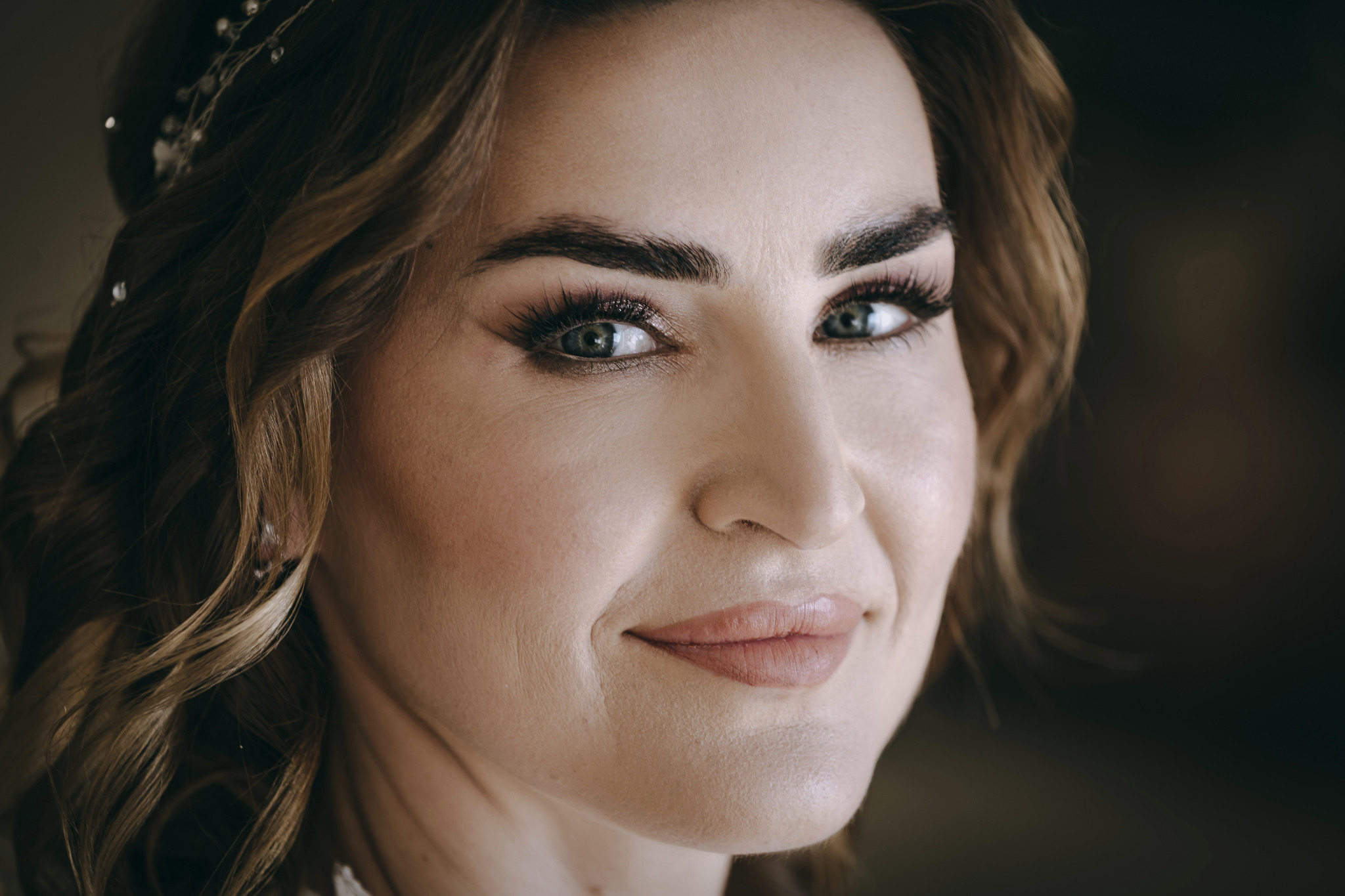 Portret kobiety z delikatnym makijażem i ozdobą we włosach, uśmiechająca się, z bliska, z ostrością na oczy i subtelne światło.
