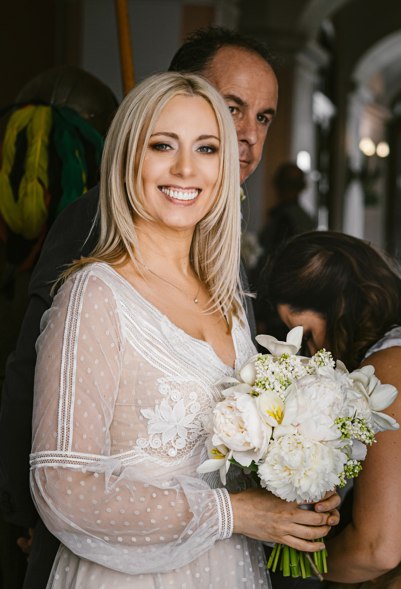Uśmiechnięta panna młoda w koronkowej sukni, trzymająca bukiet białych kwiatów, z mężczyzną w tle. Delikatne światło podkreśla detale sukni i makijaż.