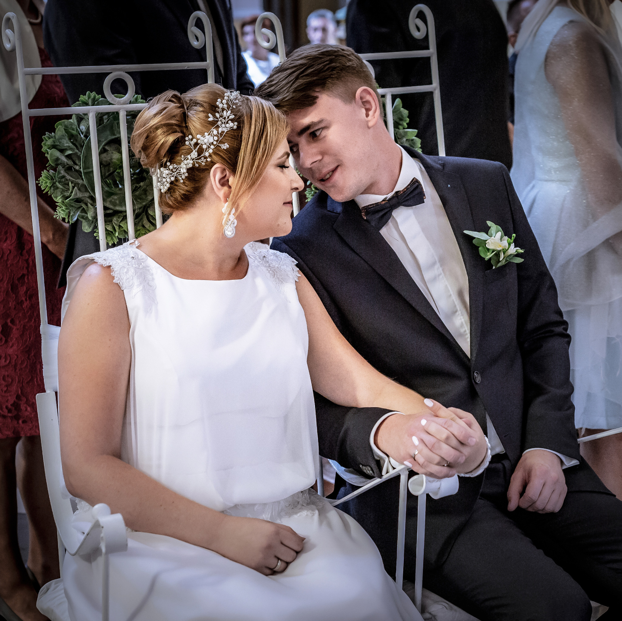 Uśmiechnięta para młoda siedzi na białym krześle, trzymając się za ręce. Panna młoda w białej sukni i ozdobie we włosach, pan młody w ciemnym garniturze z muszką.