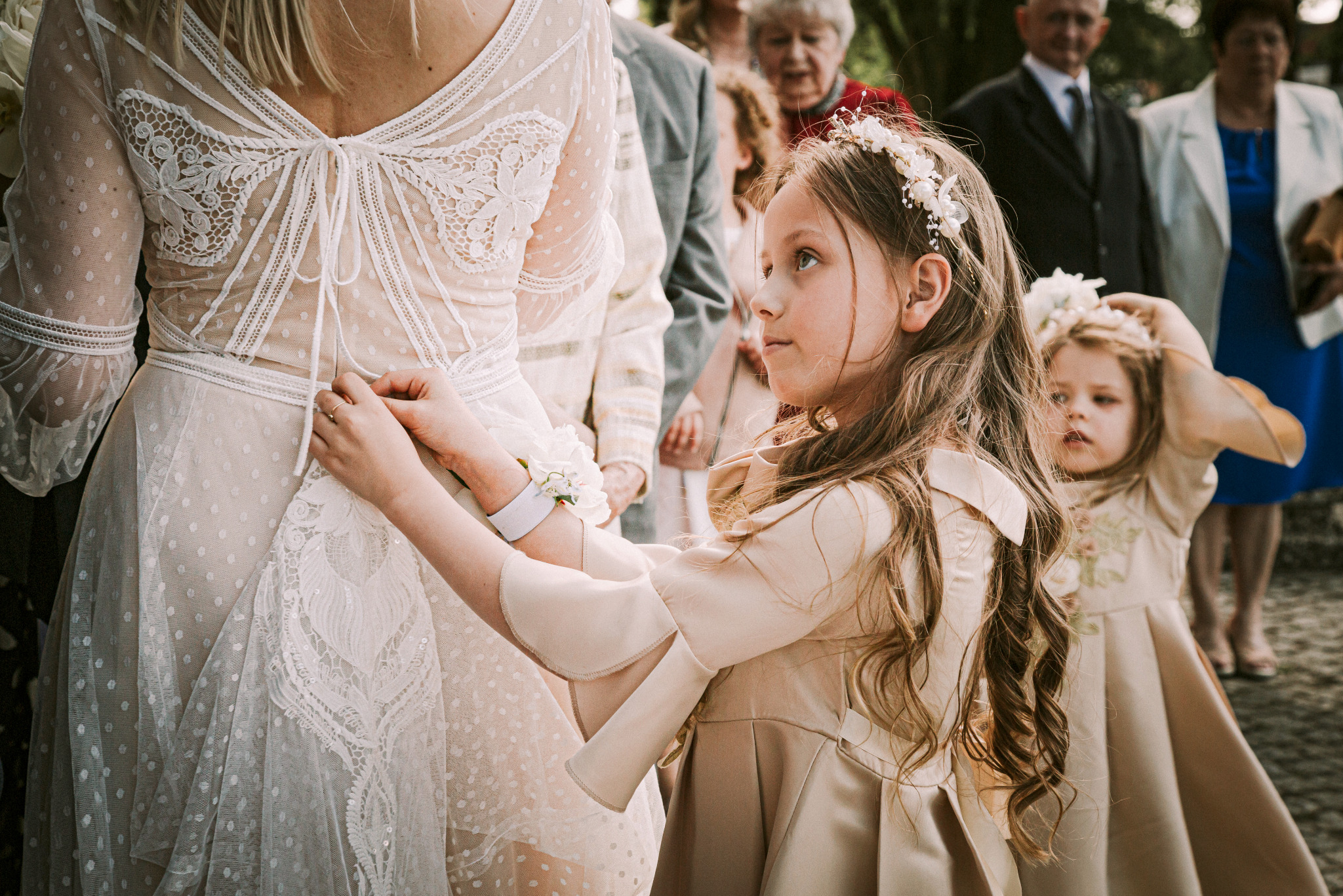 Dziewczynka w beżowej sukience poprawia wiązanie sukni ślubnej panny młodej. W tle goście weselni i druga dziewczynka w podobnej kreacji z wiankiem na głowie.