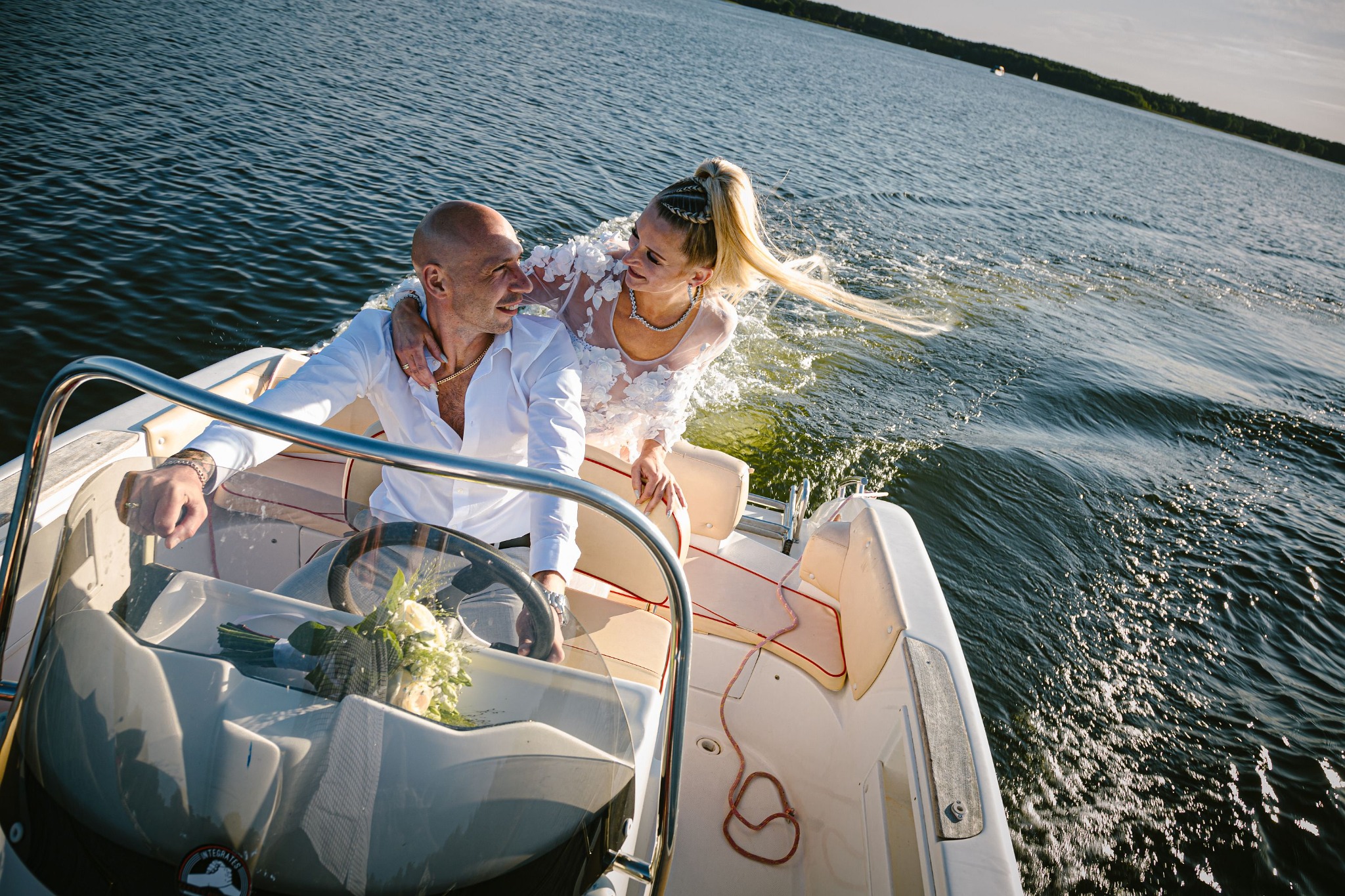 Para młoda na motorówce płynącej po jeziorze; panna młoda w białej sukni z kwiatowymi aplikacjami, z wysokim kucykiem, obejmuje pana młodego, który prowadzi łódź; bukiet ślubny leży na desce...