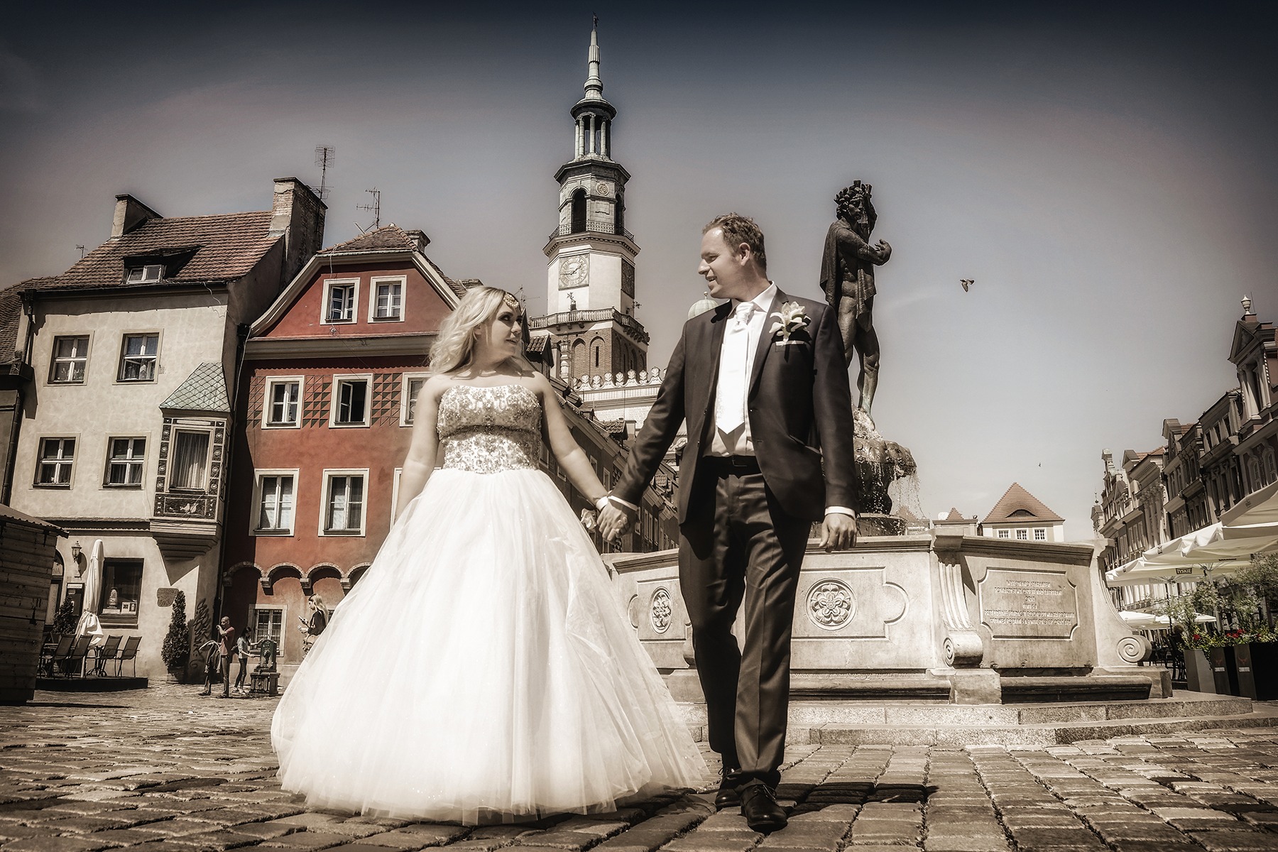 Para młoda trzymająca się za ręce na tle poznańskiego Starego Rynku, z widoczną wieżą ratuszową i fontanną z figurą Apolla, ujęcie w sepii.