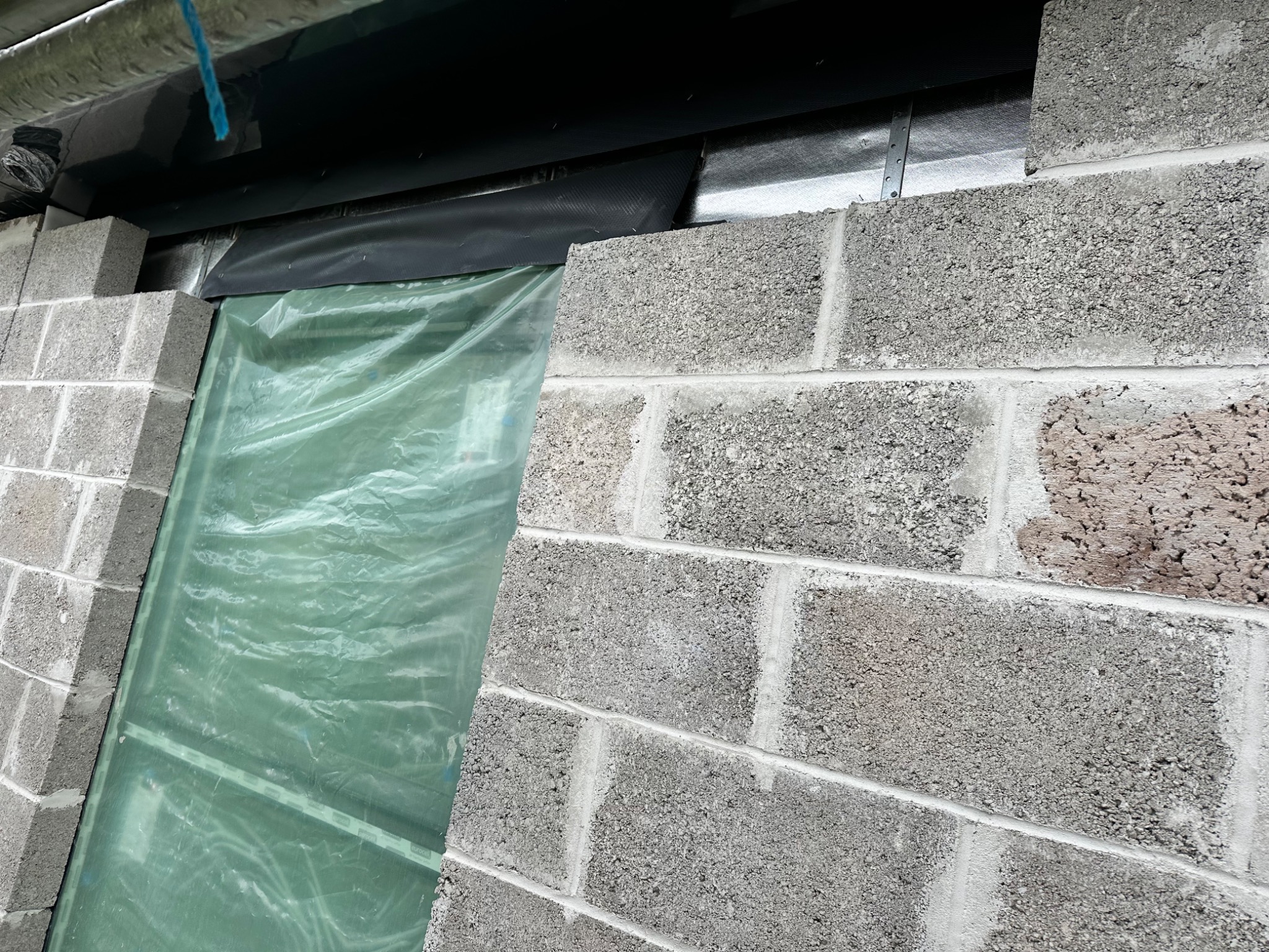 Szara ściana z bloczków betonowych w trakcie wznoszenia, zabezpieczona folią ochronną w miejscu planowanego otworu okiennego lub drzwiowego. Widoczne ślady zaprawy i nierówności powierzchni.