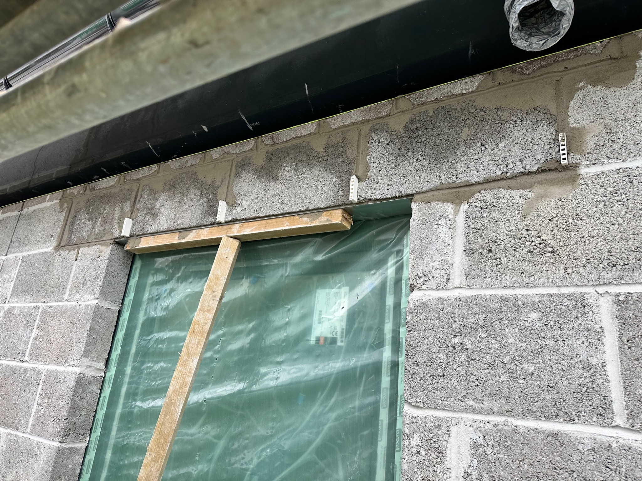 Murowanie ściany z szarego bloczka betonowego z widocznymi spoinami i fragmentem rury wentylacyjnej pod sufitem, okno zabezpieczone folią i drewnianą konstrukcją, widoczne metalowe elementy mocujące.