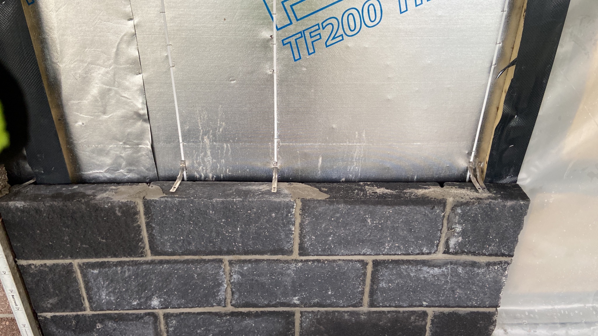 Fragment ściany z ciemnej kostki betonowej, z widoczną izolacją przeciwwilgociową TF200 i metalowymi elementami mocującymi, w trakcie montażu.