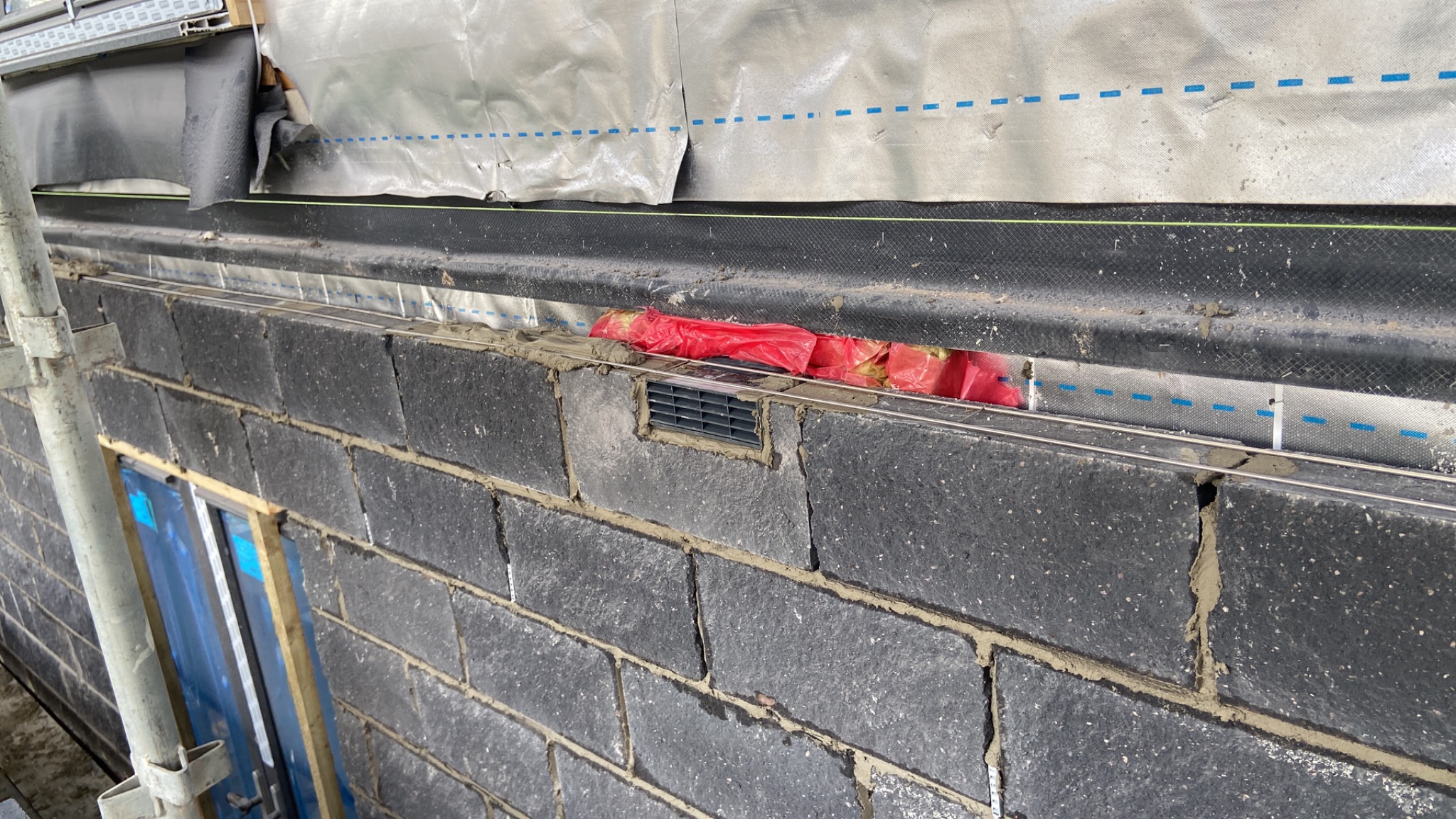 Murowana ściana z szarych bloczków, widoczna wentylacja, folia izolacyjna i fragment rusztowania z boku.