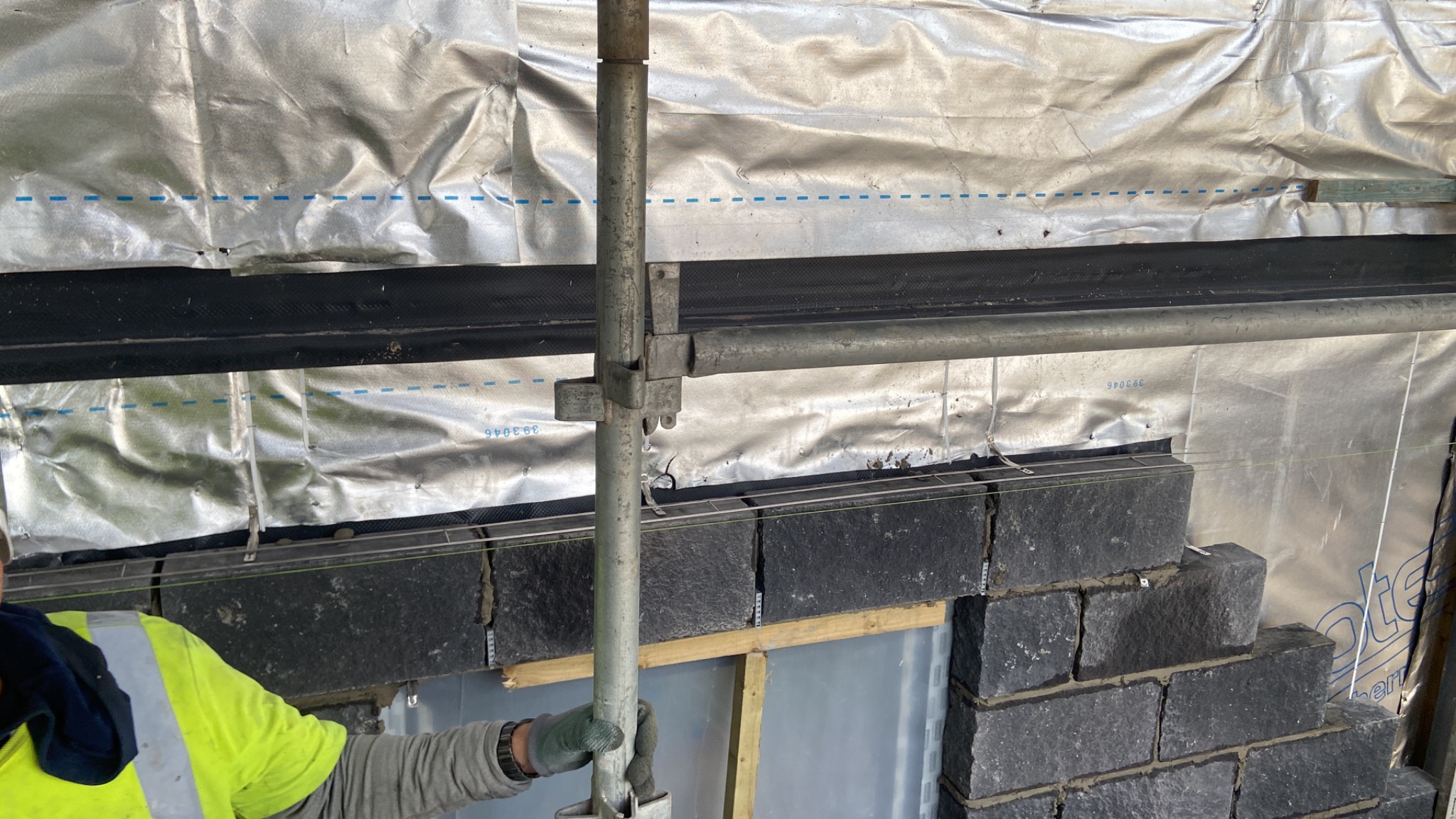 Wznoszenie ściany z ciemnych, nieregularnych kamieni, widoczne rusztowanie i izolacja przeciwwilgociowa, pracownik w odblaskowej kamizelce muruje.