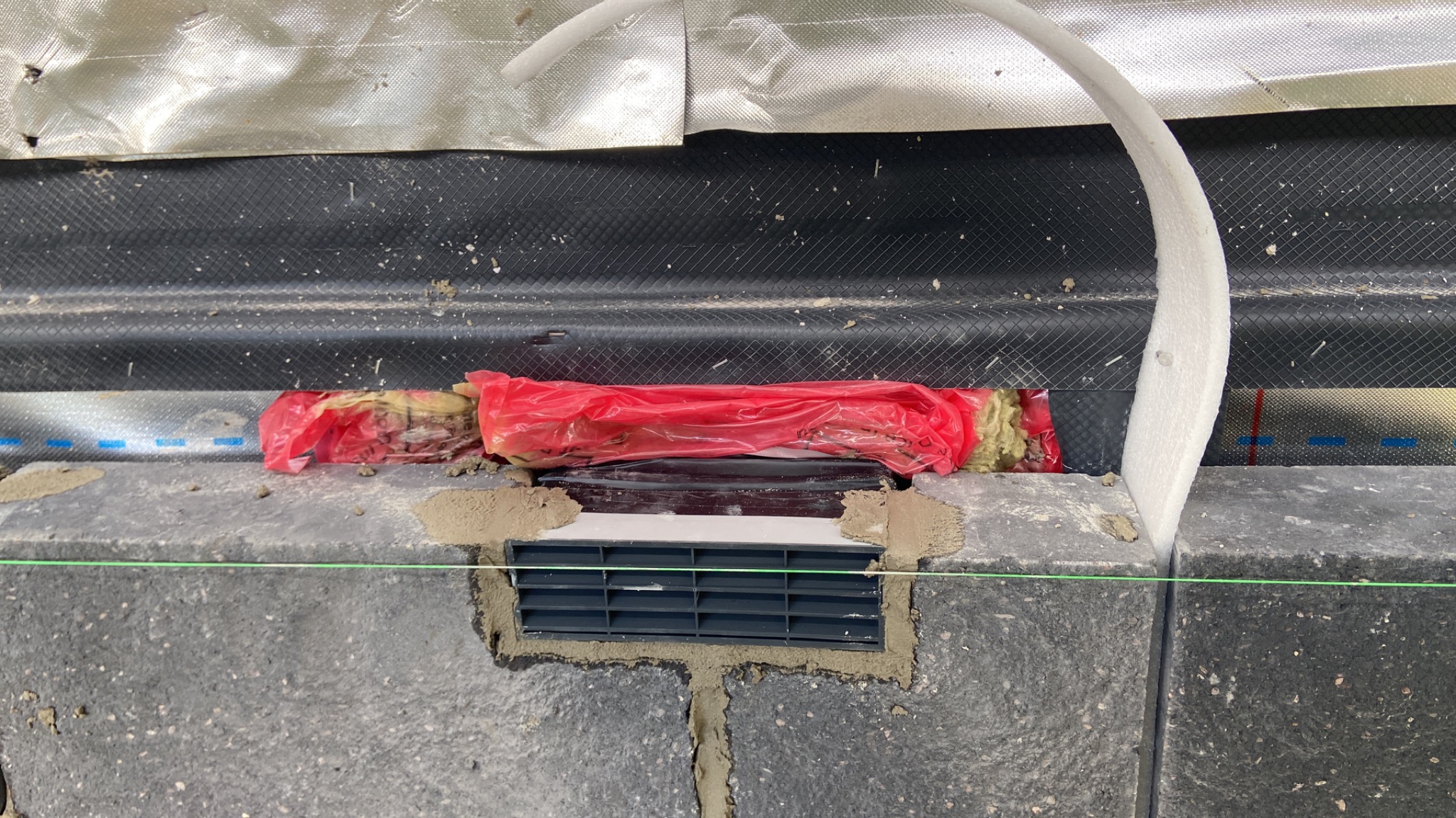 Szczegółowe ujęcie fragmentu elewacji z zamontowaną kratką wentylacyjną, zabezpieczoną czerwoną folią i wełną mineralną, z widoczną warstwą hydroizolacji i przewodem instalacyjnym.