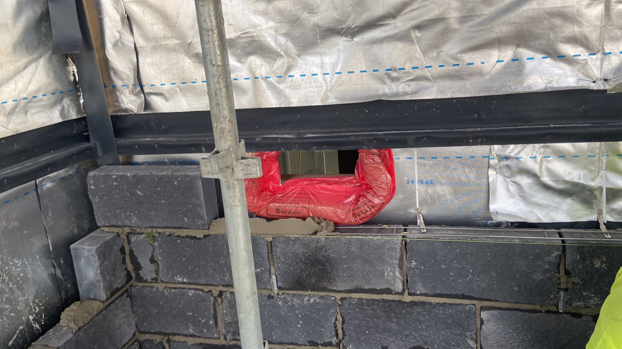 Wznoszenie ściany z ciemnych bloczków betonowych, z widocznym rusztowaniem i zabezpieczeniem otworu okiennego czerwoną folią.