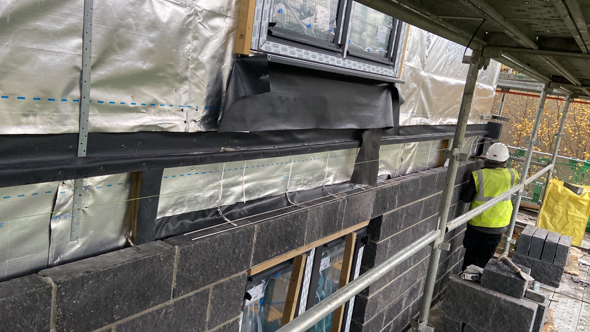 Wznoszenie ściany z ciemnych bloczków betonowych z widoczną izolacją termiczną i folią paroizolacyjną, pracownik w kasku i kamizelce na rusztowaniu, widoczne okna w trakcie montażu.