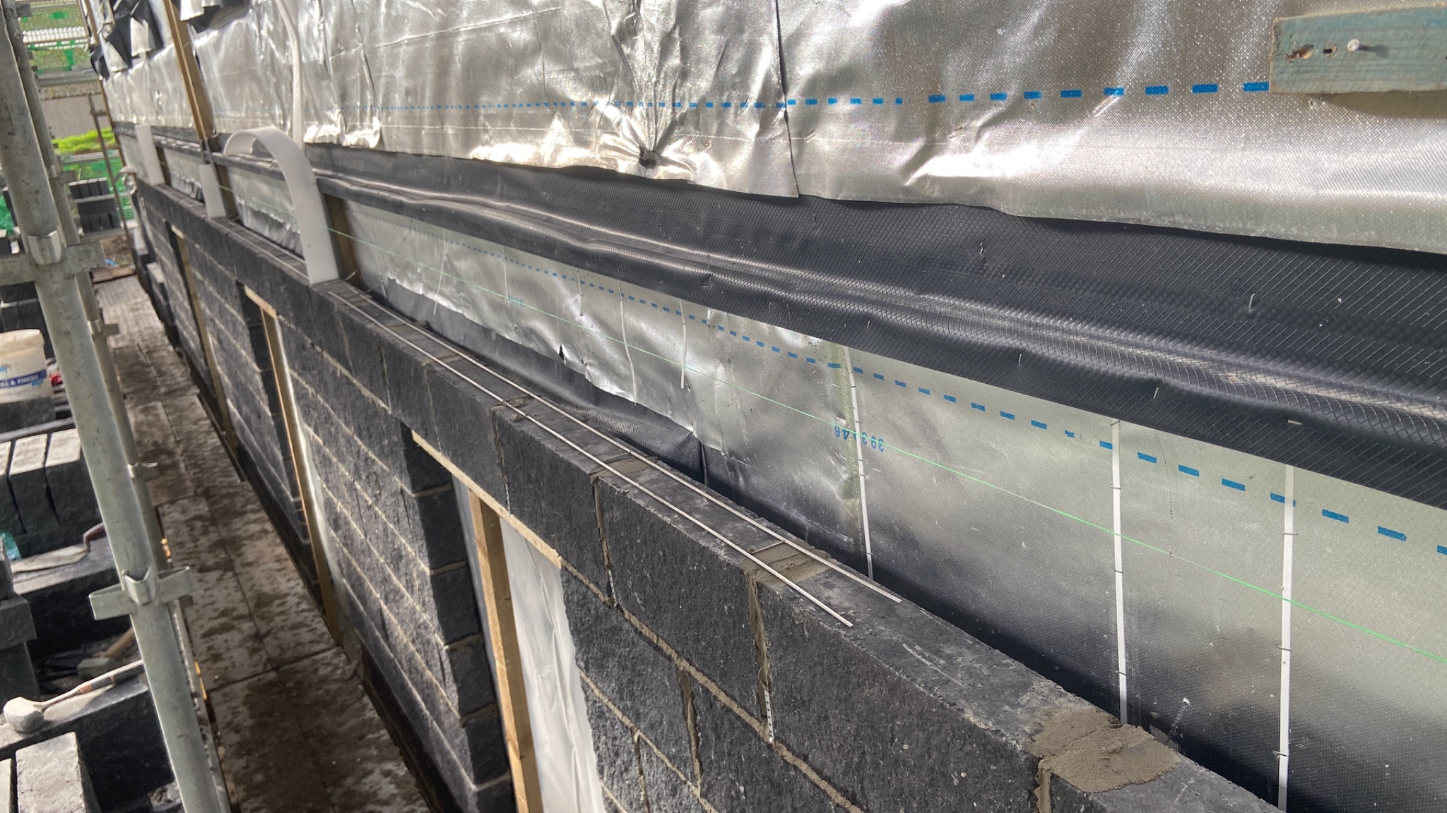 Wznoszenie ściany z ciemnoszarych bloczków betonowych, widoczne zbrojenie poziome, izolacja przeciwwilgociowa i rusztowanie w tle.