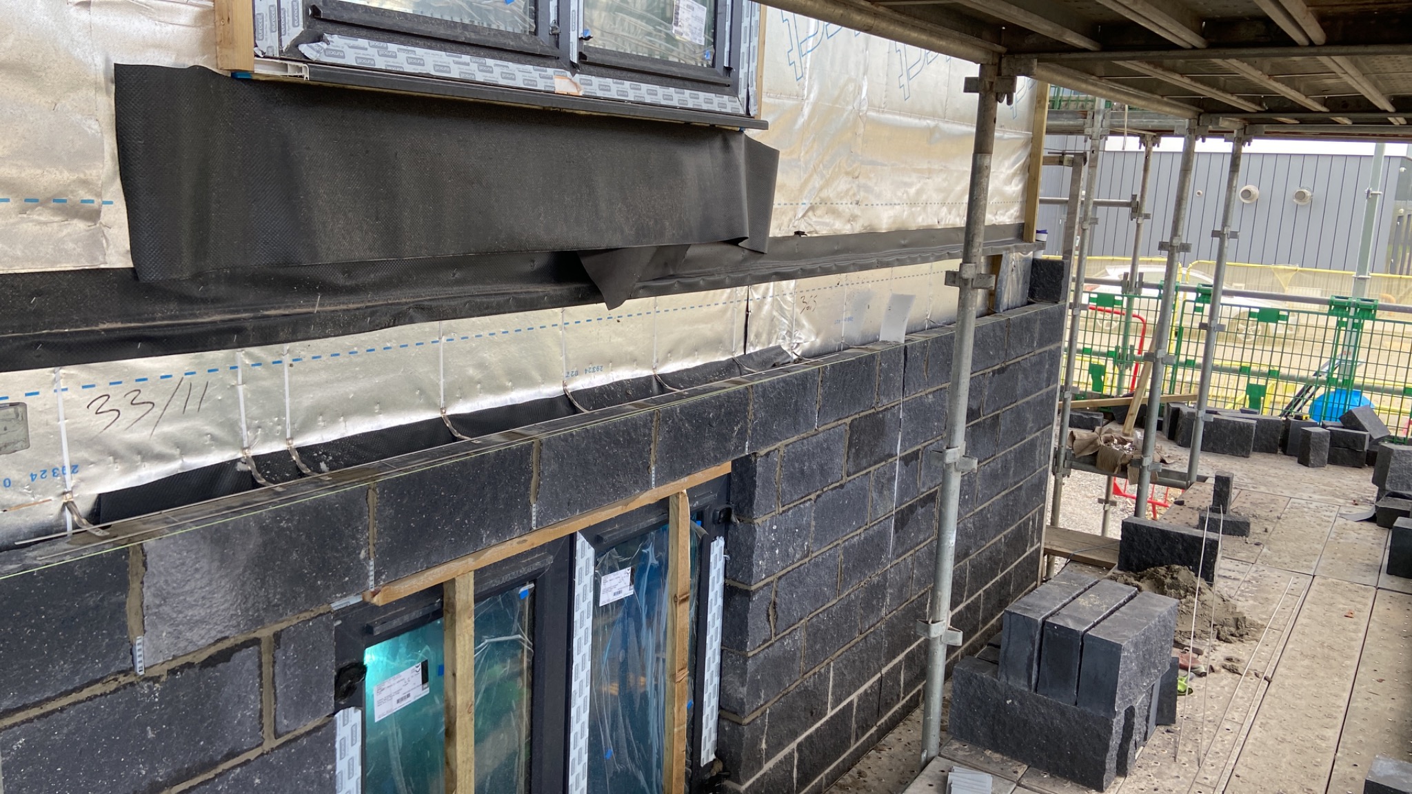 Wznoszenie muru z czarnego kamienia elewacyjnego na tle rusztowania i okien z folią ochronną, widoczne bloczki kamienne, izolacja termiczna na ścianie.