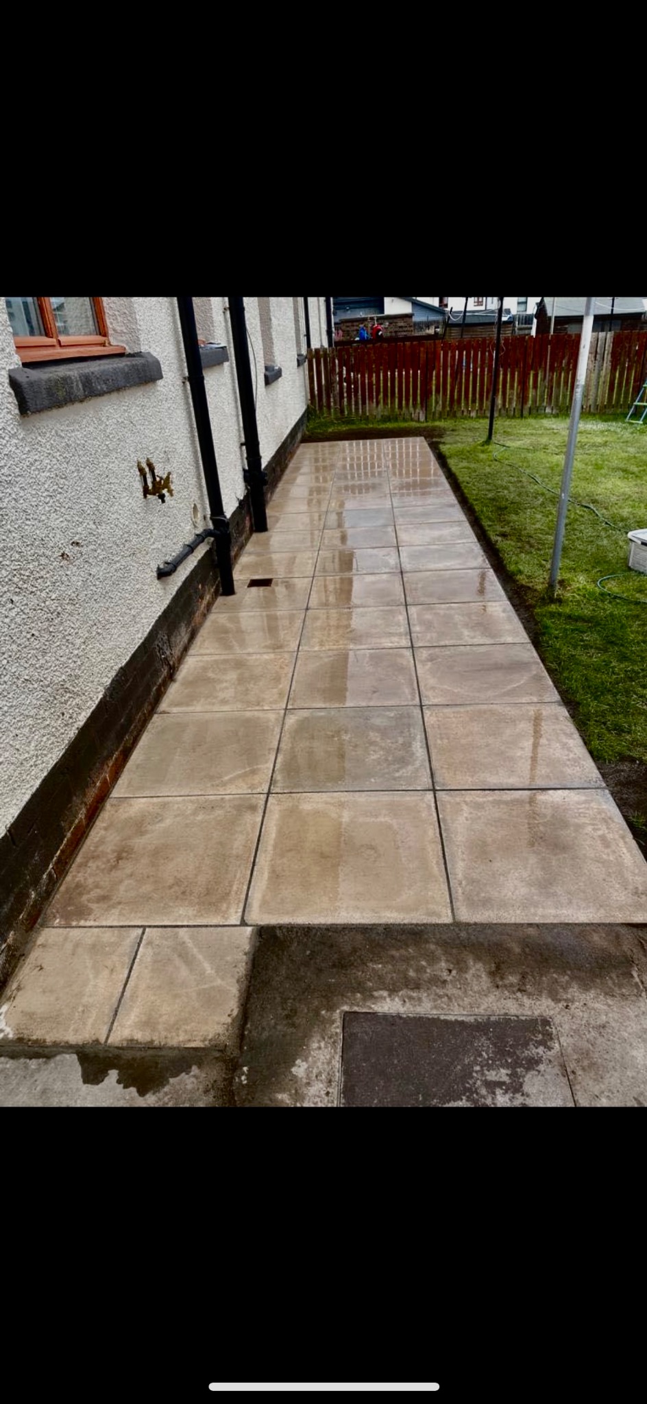 Ułożona kostka brukowa przy ścianie domu, widoczny fragment trawnika i ogrodzenia, świeżo po deszczu, mokra powierzchnia kostki odbija światło.