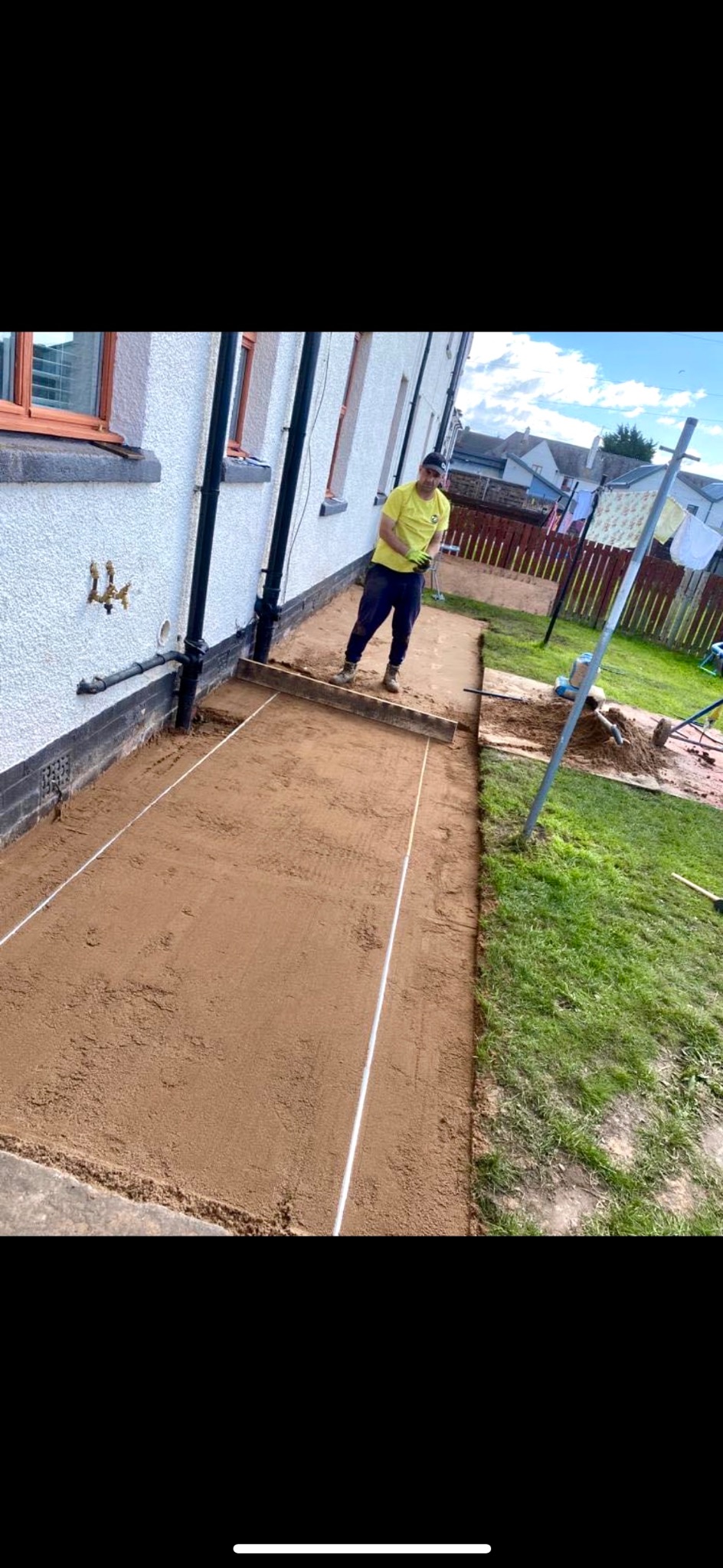 Przygotowanie podłoża pod ścieżkę z kostki brukowej: wyrównany piasek ograniczony białymi linkami, pracownik w żółtej koszulce i rękawicach roboczych, widoczny fragment ściany domu i trawnika w tle.