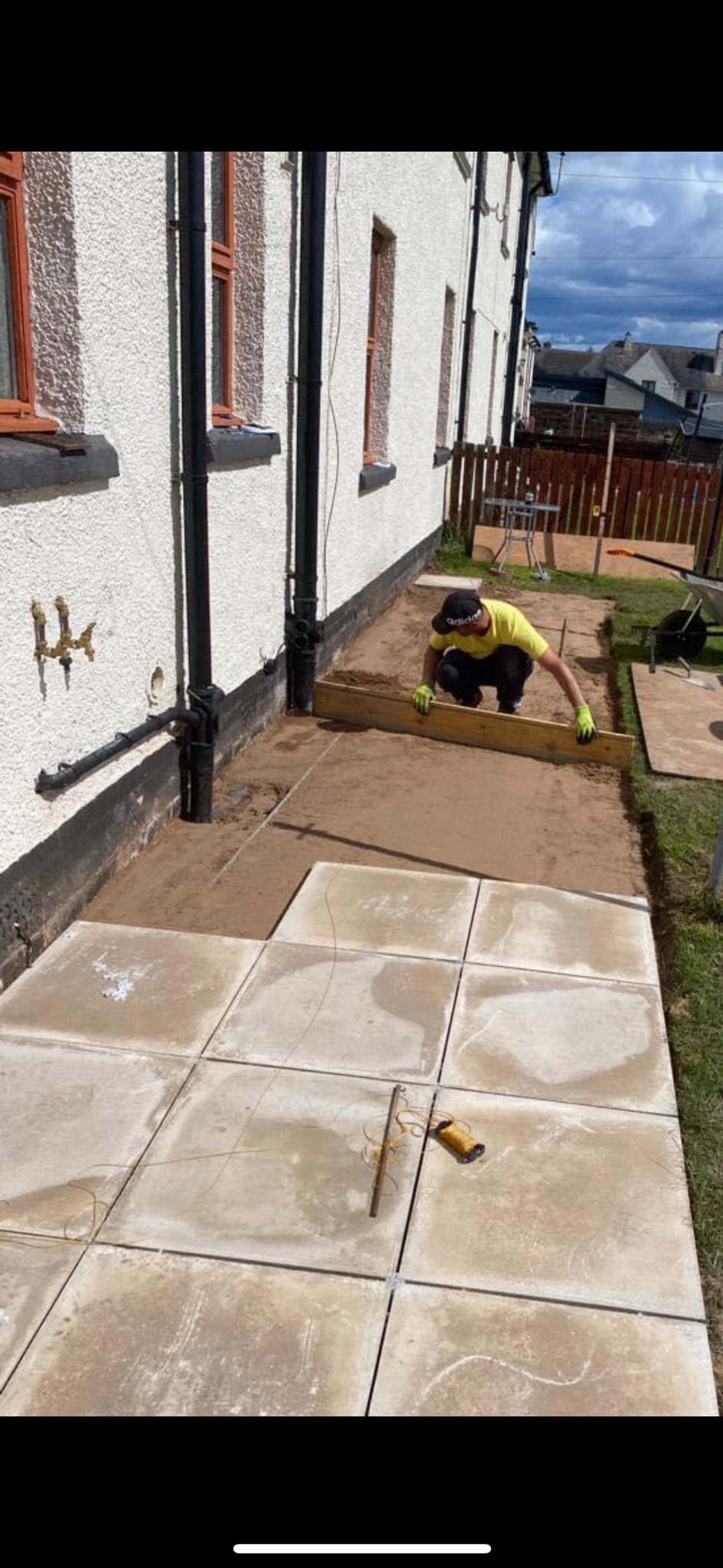 Pracownik w żółtej koszulce i rękawicach wyrównuje piasek pod płyty chodnikowe przy białym budynku z brązowymi oknami, widoczne narzędzia i sznurek do wyznaczania linii.