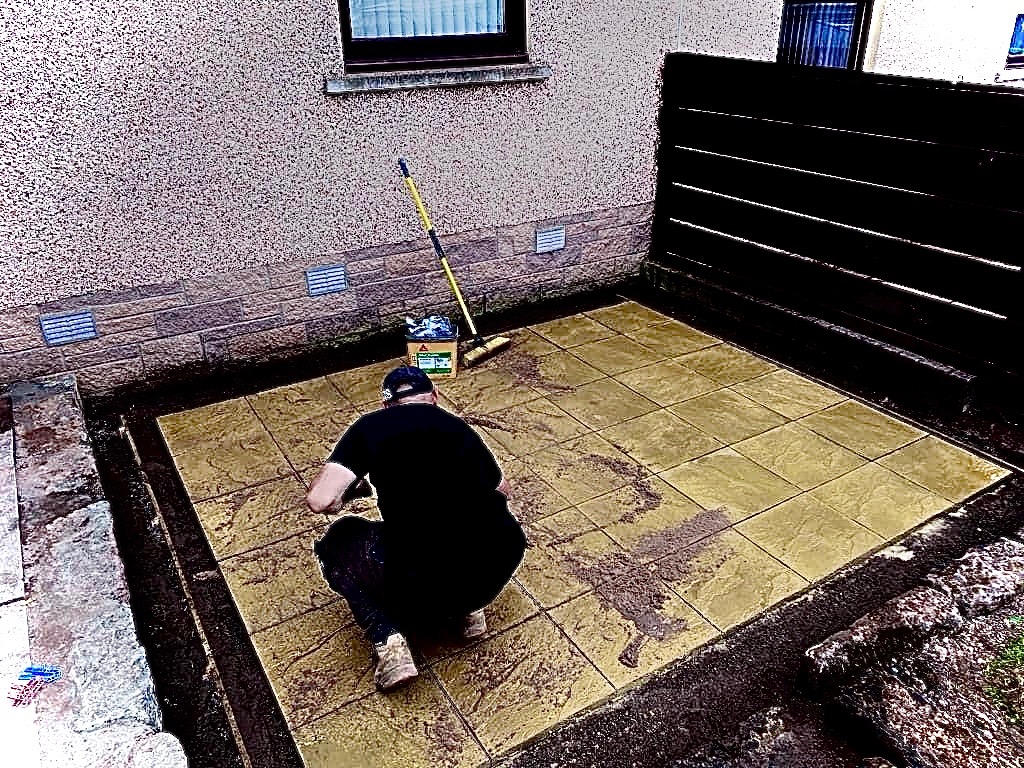 Pracownik na kolanach czyści nowo ułożoną, kwadratową, żółtą kostkę brukową na patio; widoczna szczotka i pojemnik z preparatem czyszczącym, ciemne ogrodzenie w tle.