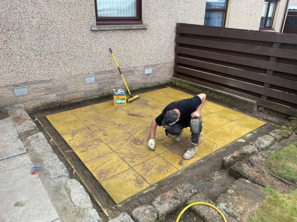 Pracownik w rękawicach i nakolannikach układa żółtą kostkę brukową na przygotowanym podłożu, widoczna szczotka i pojemnik z materiałem do fugowania, obrzeże z kamieni i drewniany płot w tle.
