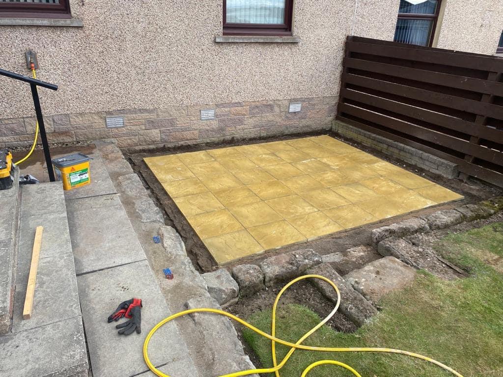 Żółta kostka brukowa ułożona na przygotowanym podłożu, widoczne narzędzia, węże i rękawice robocze, w tle ogrodzenie z desek i elewacja budynku.