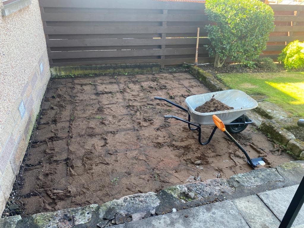 Przygotowanie podłoża pod nawierzchnię: wyrównany teren z widocznymi śladami po kostce, taczka z ziemią i łopata z pomarańczową rączką, drewniany płot w tle.