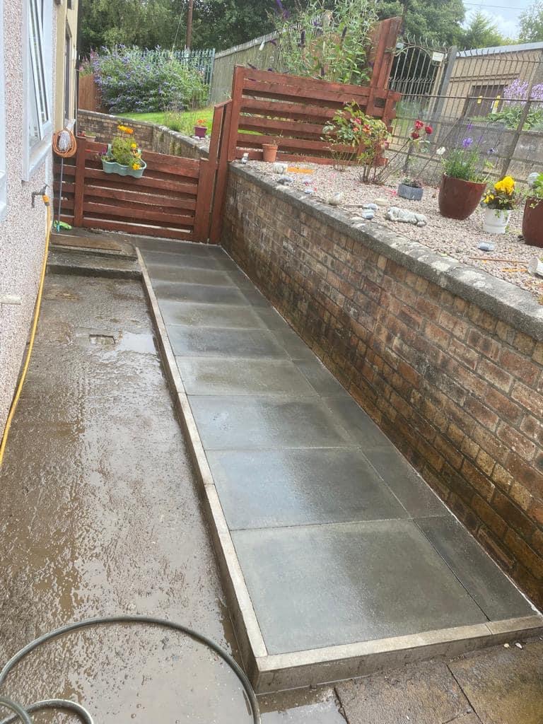 Świeżo ułożony, mokry chodnik z szarych płyt betonowych prowadzący do drewnianej furtki w ogrodzeniu, z widocznymi roślinami w donicach na murku z cegły.