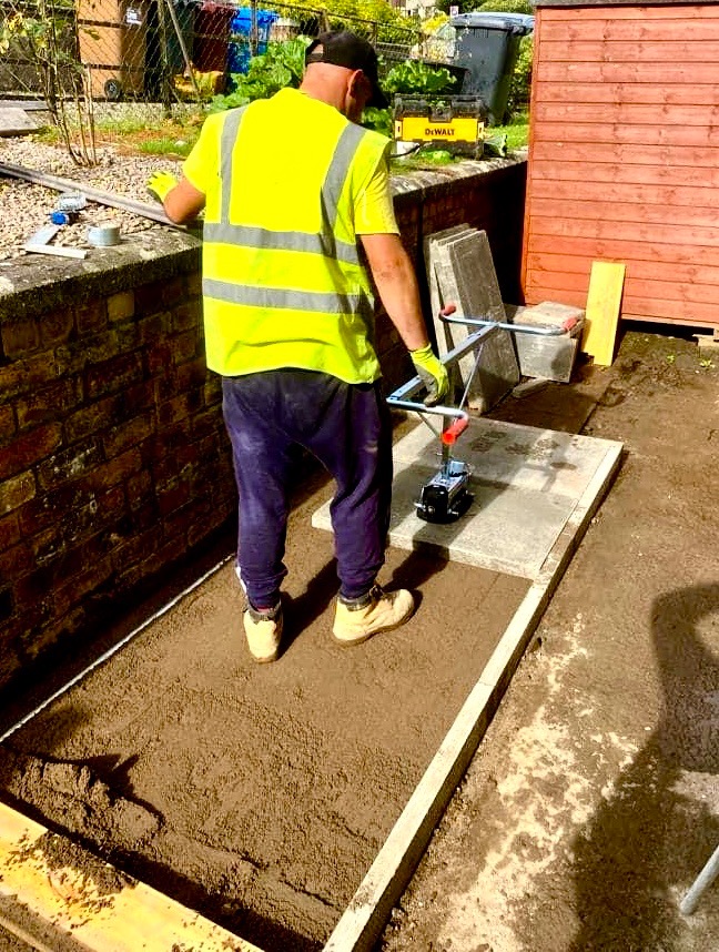 Pracownik w żółtej kamizelce i roboczych butach układa betonowe płyty za pomocą specjalnego podnośnika próżniowego na przygotowanym podłożu z piasku, obok widoczny mur z cegły i narzędzia.