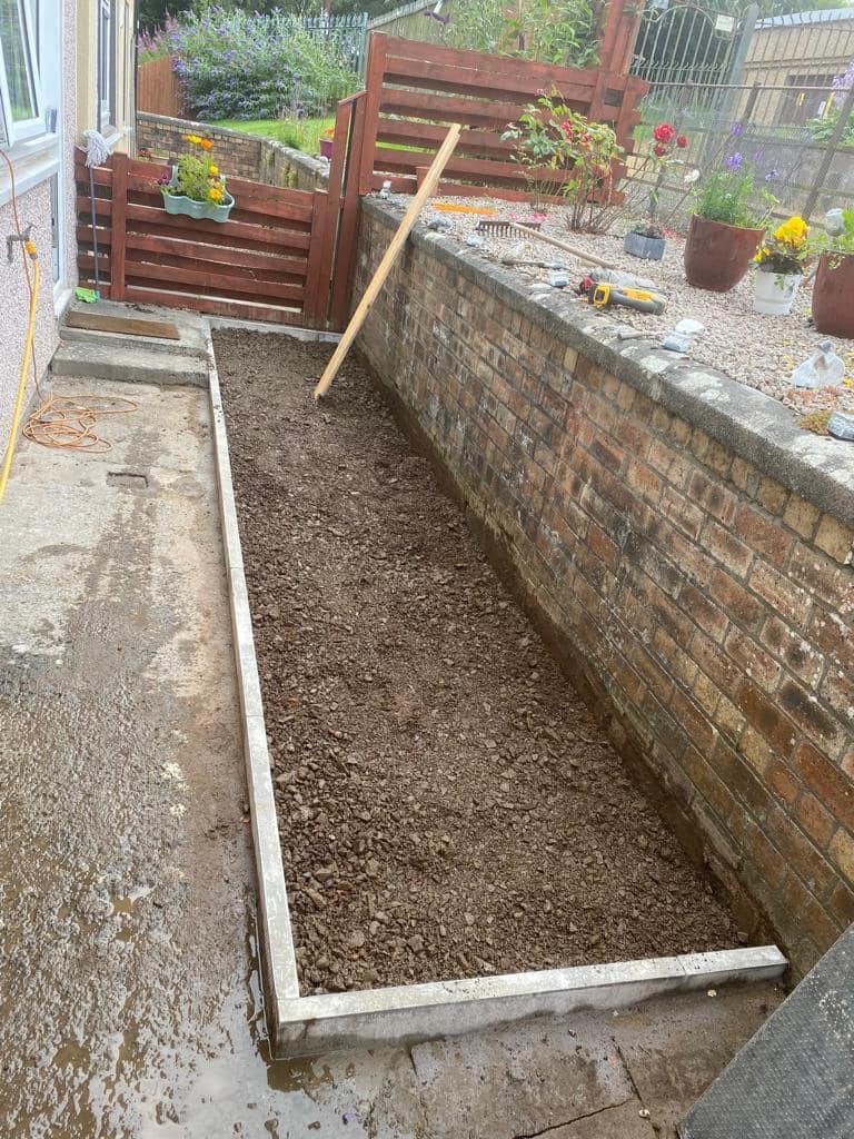 Przygotowany teren pod ścieżkę ogrodową z obrzeżami, ubity gruz i narzędzia ogrodnicze w tle.