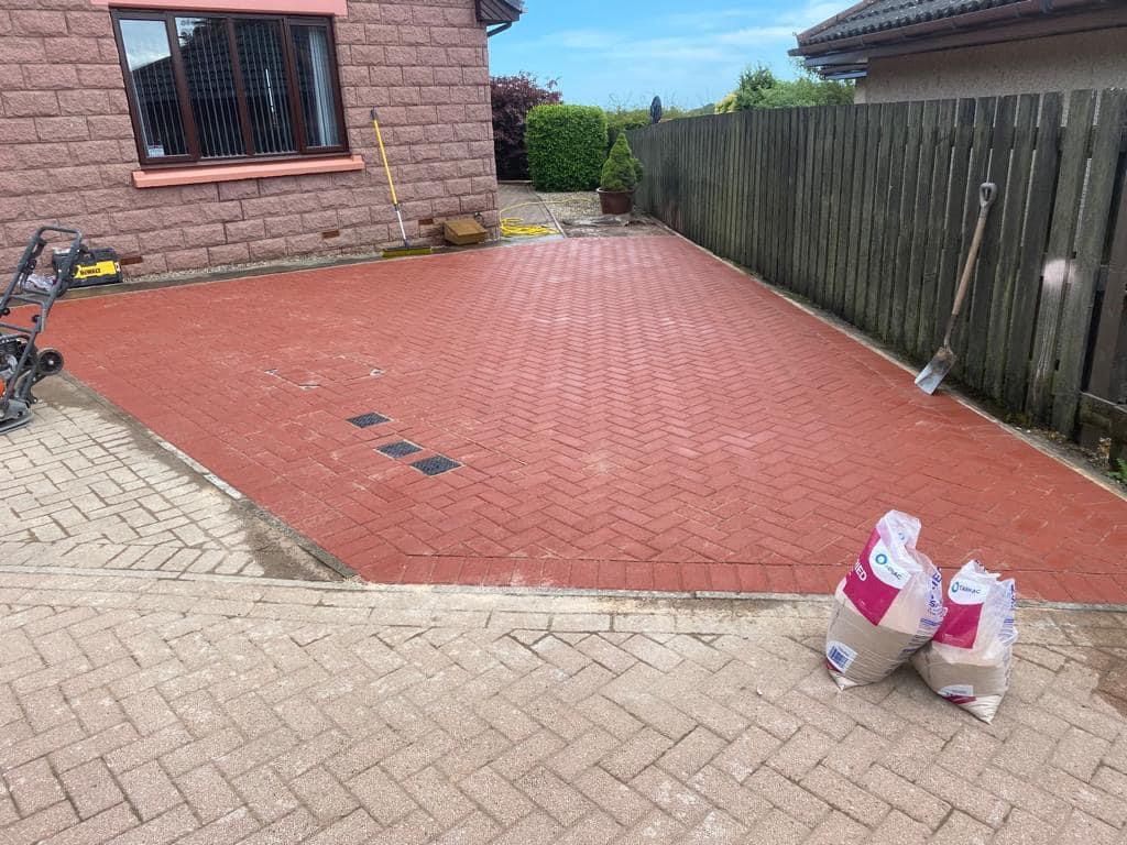 Czerwona kostka brukowa ułożona w jodełkę na podjeździe, z widocznym zagęszczarką, workami piasku do spoinowania i narzędziami obok drewnianego ogrodzenia.