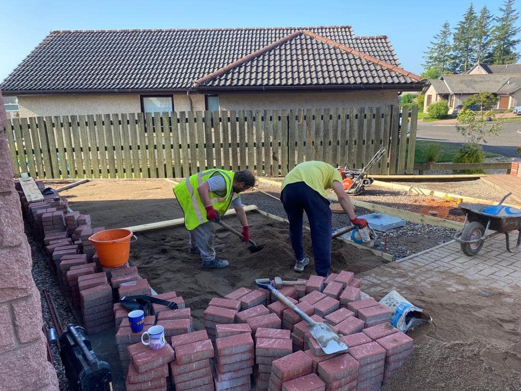 Dwóch robotników w odblaskowych kamizelkach przygotowuje podłoże pod ułożenie czerwonej kostki brukowej na posesji przed domem jednorodzinnym. Na pierwszym planie stosy kostki, kubki i narzędzia.