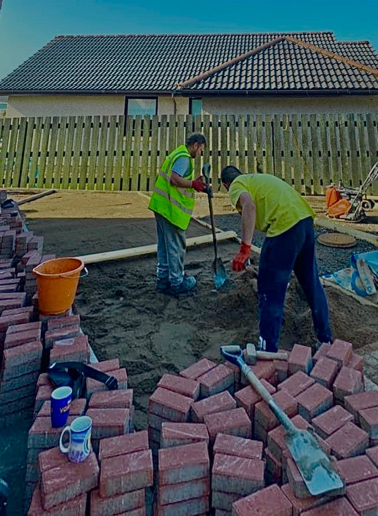 Dwóch pracowników w odblaskowych kamizelkach przygotowuje podłoże pod ułożenie czerwonej kostki brukowej, na pierwszym planie stosy kostki, narzędzia i kubki.