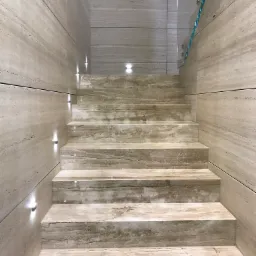 Schody wewnętrzne wyłożone trawertynem z dyskretnym oświetleniem punktowym w ścianie i szklaną balustradą.