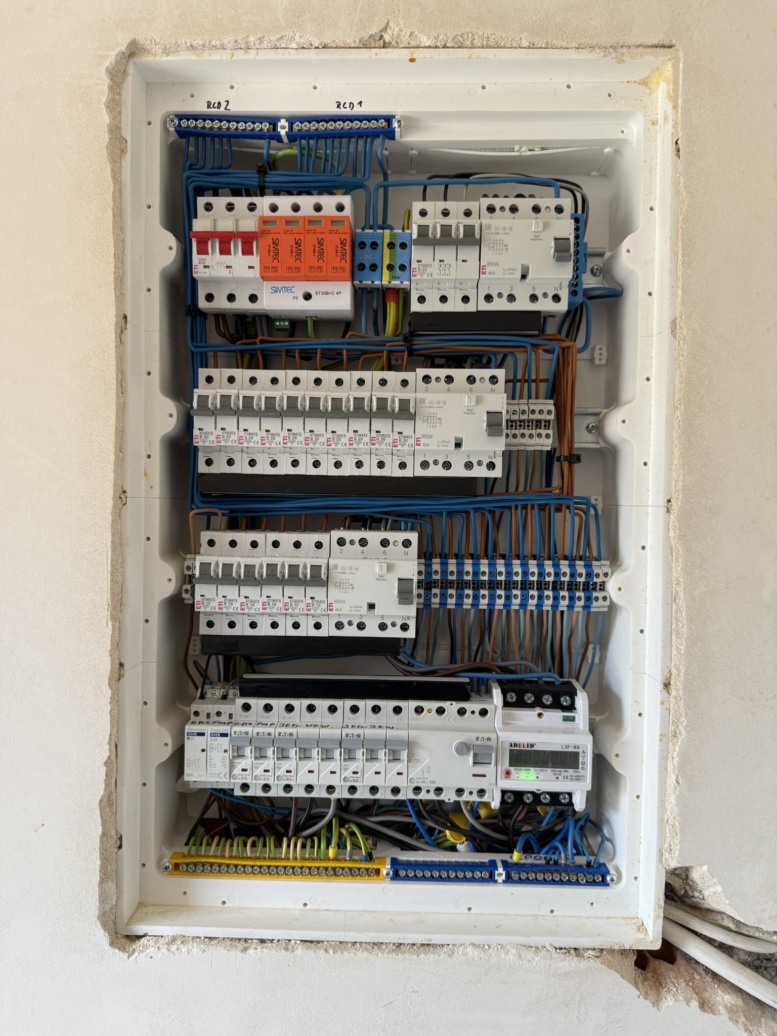 Rozdzielnica elektryczna w białej obudowie, z widocznymi bezpiecznikami, przewodami i elementami montażowymi. Instalacja w surowym stanie, przed wykończeniem ściany.