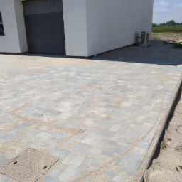 Szara kostka brukowa na podjeździe domu jednorodzinnego, z widoczną studzienką kanalizacyjną i piaskiem wypełniającym szczeliny między kostkami, budynek z garażem w tle.