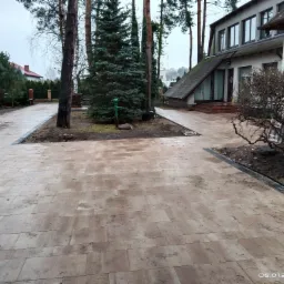 Nowa nawierzchnia z jasnych, prostokątnych płyt ułożona na posesji przed domem z ogrodem, widoczny fragment elewacji budynku i drzewa.