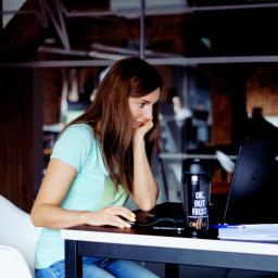 Seology sp. z o.o. - Kobieta z brązowymi włosami, w jasnej koszulce, siedzi przy biurku z laptopem i kubkiem z napisem 'OK. BUT FIRST coffee', kalendarzem wskazującym 16 lipca i dokumentami, w nowoczesnym biurze...