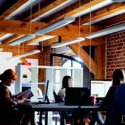 Wnętrze biura z odsłoniętą cegłą i drewnianymi belkami stropowymi; pracownicy przy biurkach z komputerami, jedna osoba czyta książkę.
