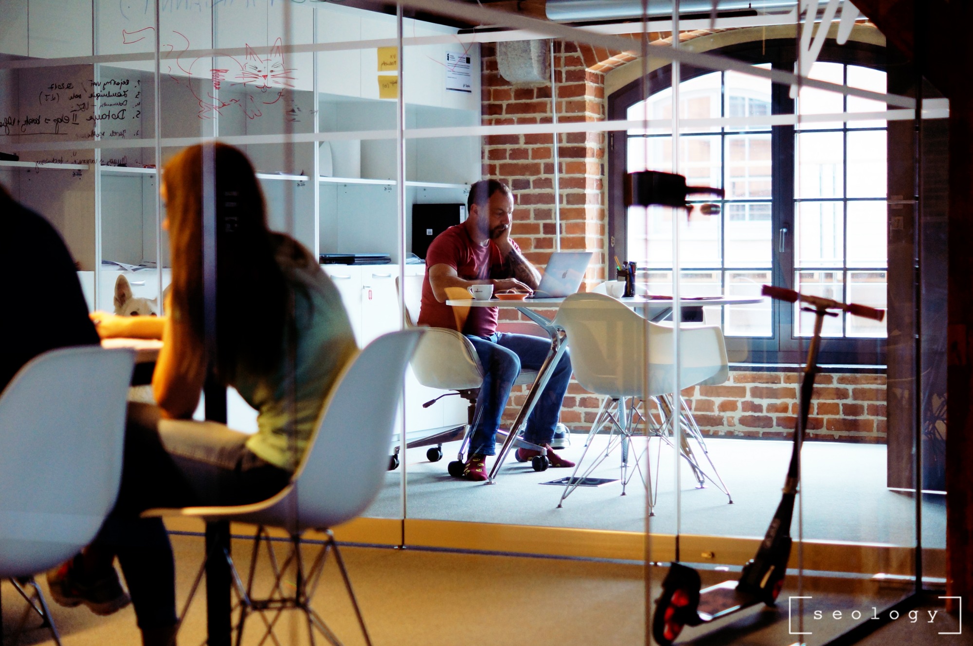 Wnętrze biura z przeszkloną ścianą, za którą widać mężczyznę pracującego na laptopie przy biurku, z filiżanką kawy i przekąskami, oraz kobietę siedzącą przy innym biurku, a w tle biała tablica...