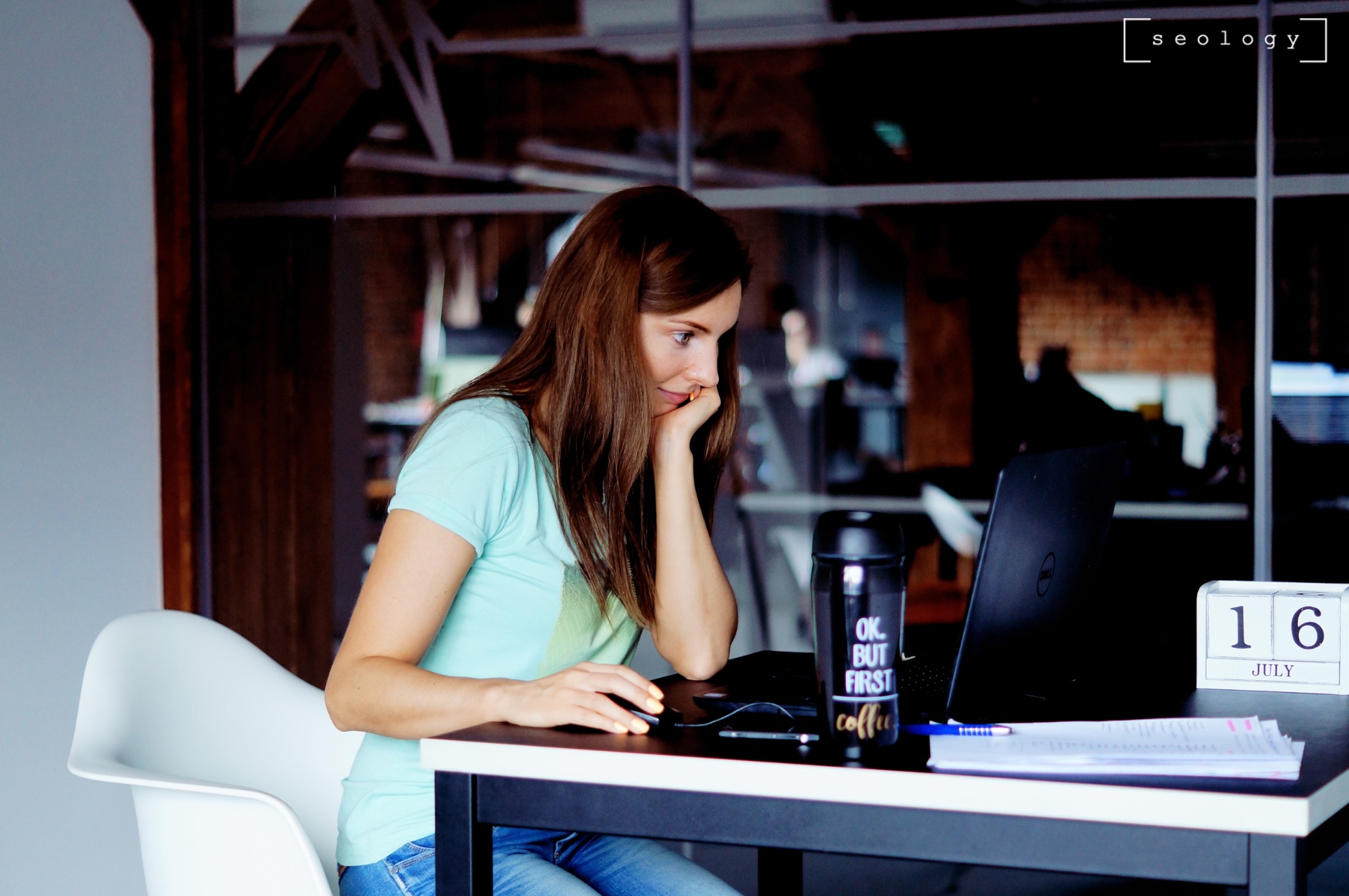 Kobieta z brązowymi włosami, w jasnej koszulce, siedzi przy biurku z laptopem i kubkiem z napisem 'OK. BUT FIRST coffee', kalendarzem wskazującym 16 lipca i dokumentami, w nowoczesnym biurze...