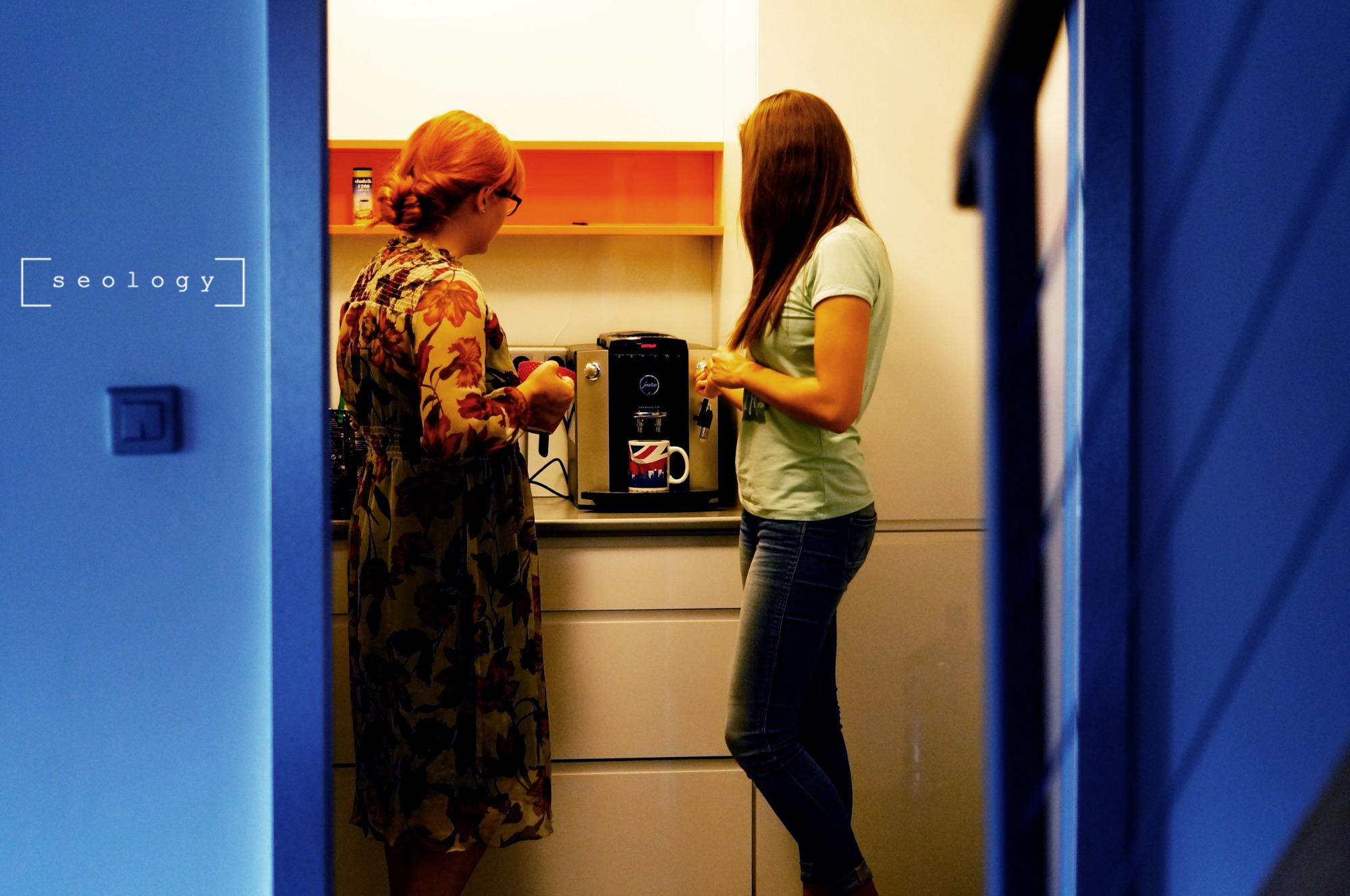 Dwie kobiety rozmawiają przy ekspresie do kawy Jura z kubkiem z flagą Wielkiej Brytanii w biurowej kuchni z pomarańczową półką, widok zza niebieskich drzwi z napisem 'seology'.