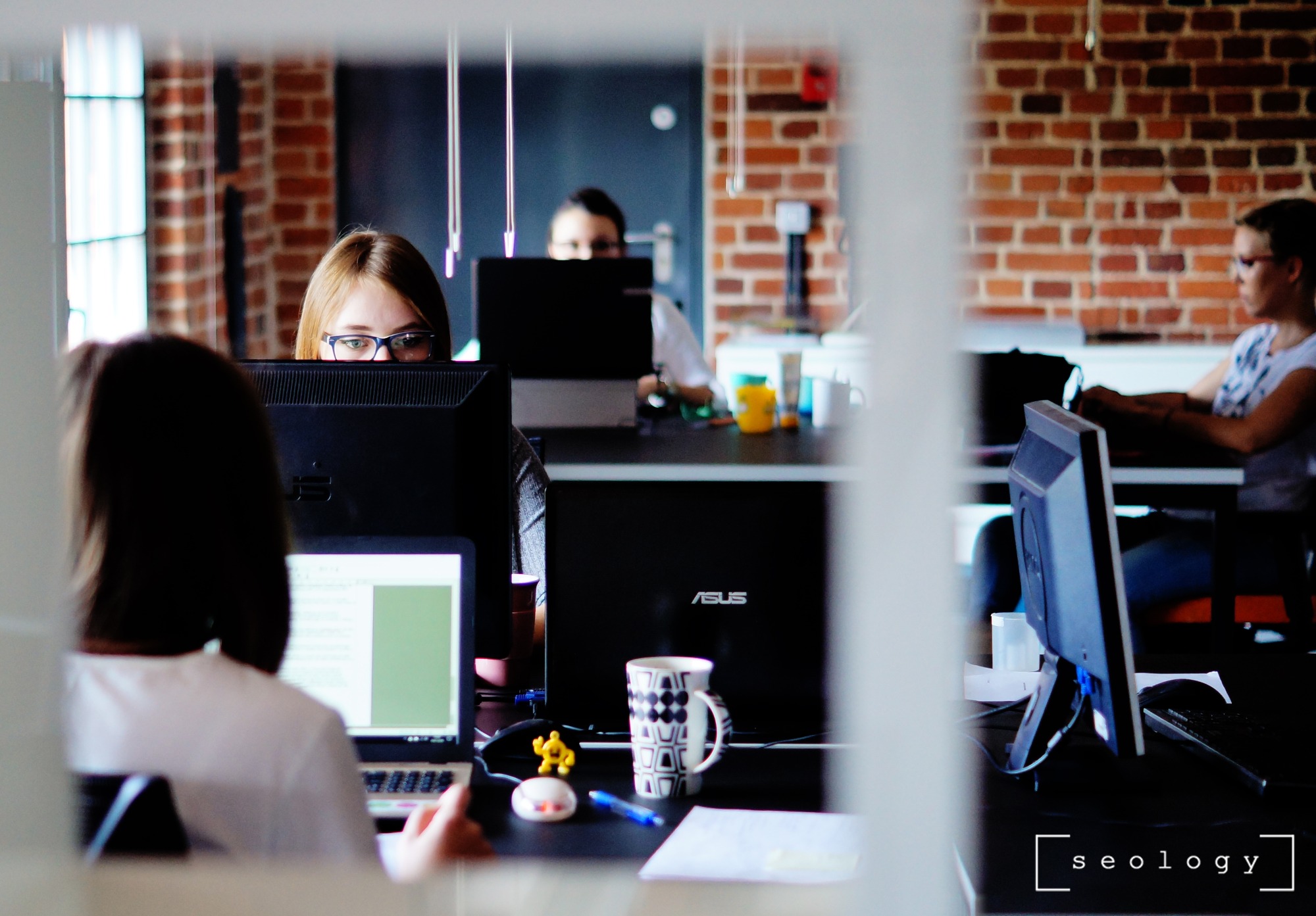 Biuro z ceglaną ścianą, kilka osób pracuje przy komputerach, na biurku kubek i figurka, widok przez szklaną ścianę.