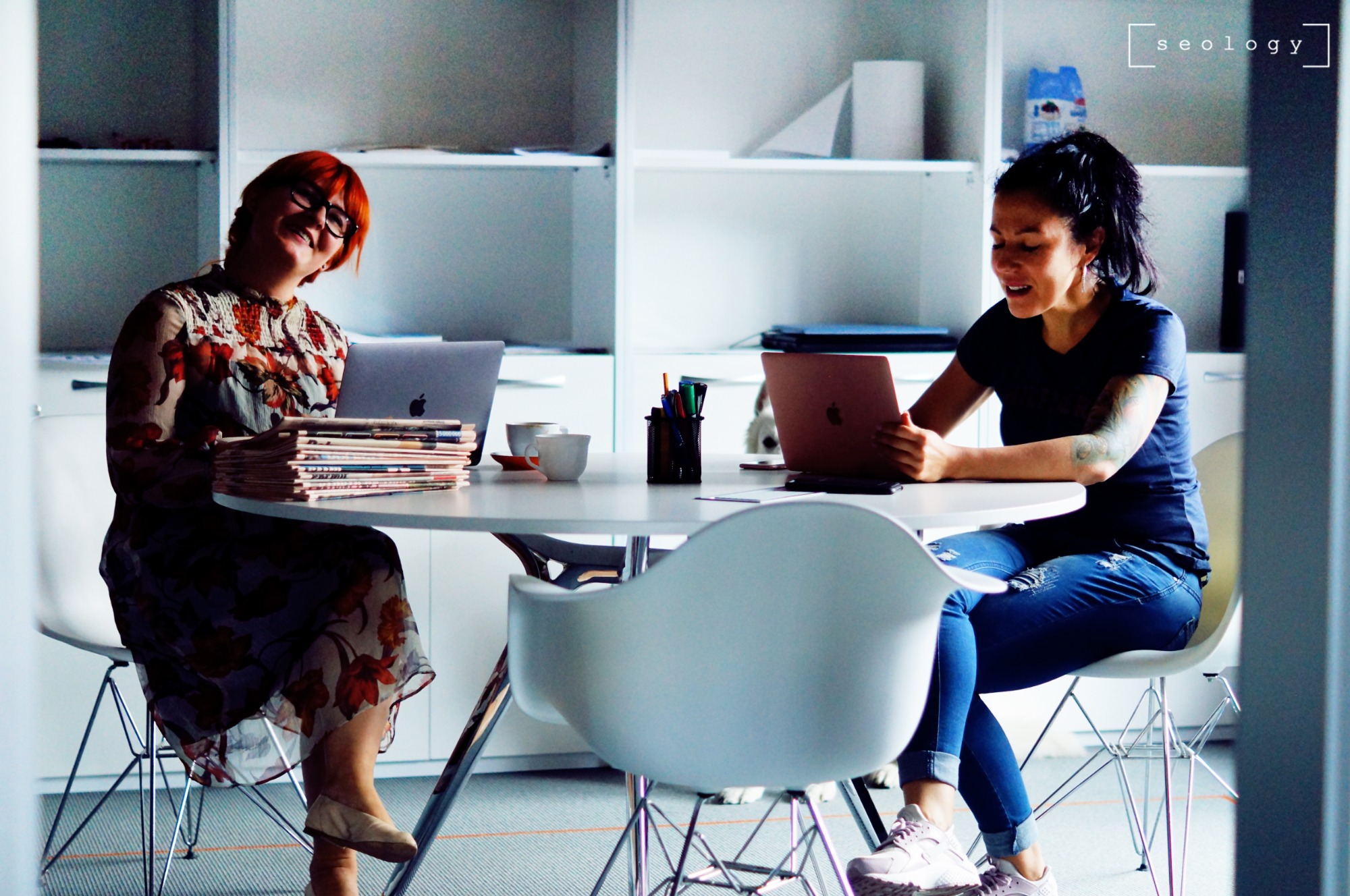 Dwie kobiety pracują przy okrągłym stole w biurze, jedna z laptopem i stosem gazet, druga z tabletem, obie uśmiechnięte.