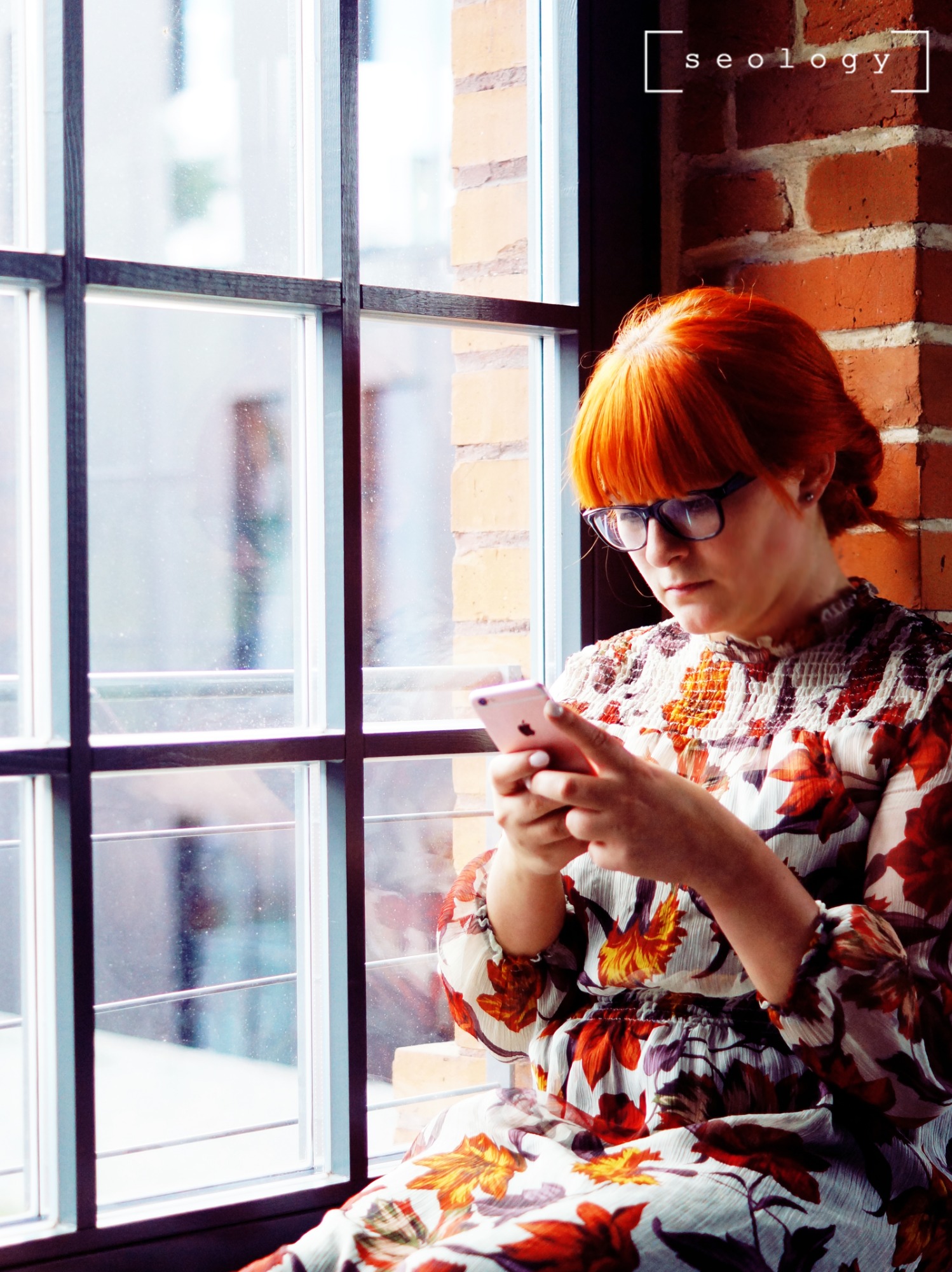 Kobieta z rudymi włosami w okularach, siedząca przy oknie z ceglaną ścianą w tle, przegląda coś na smartfonie.