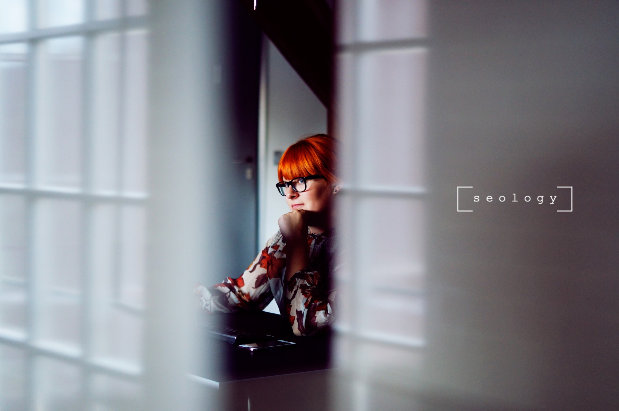 Kobieta z rudymi włosami i okularami siedzi zamyślona przy biurku, na którym leży smartfon, widoczna przez rozmyte pionowe linie, z napisem 'seology' w kwadratowych nawiasach po prawej stronie.