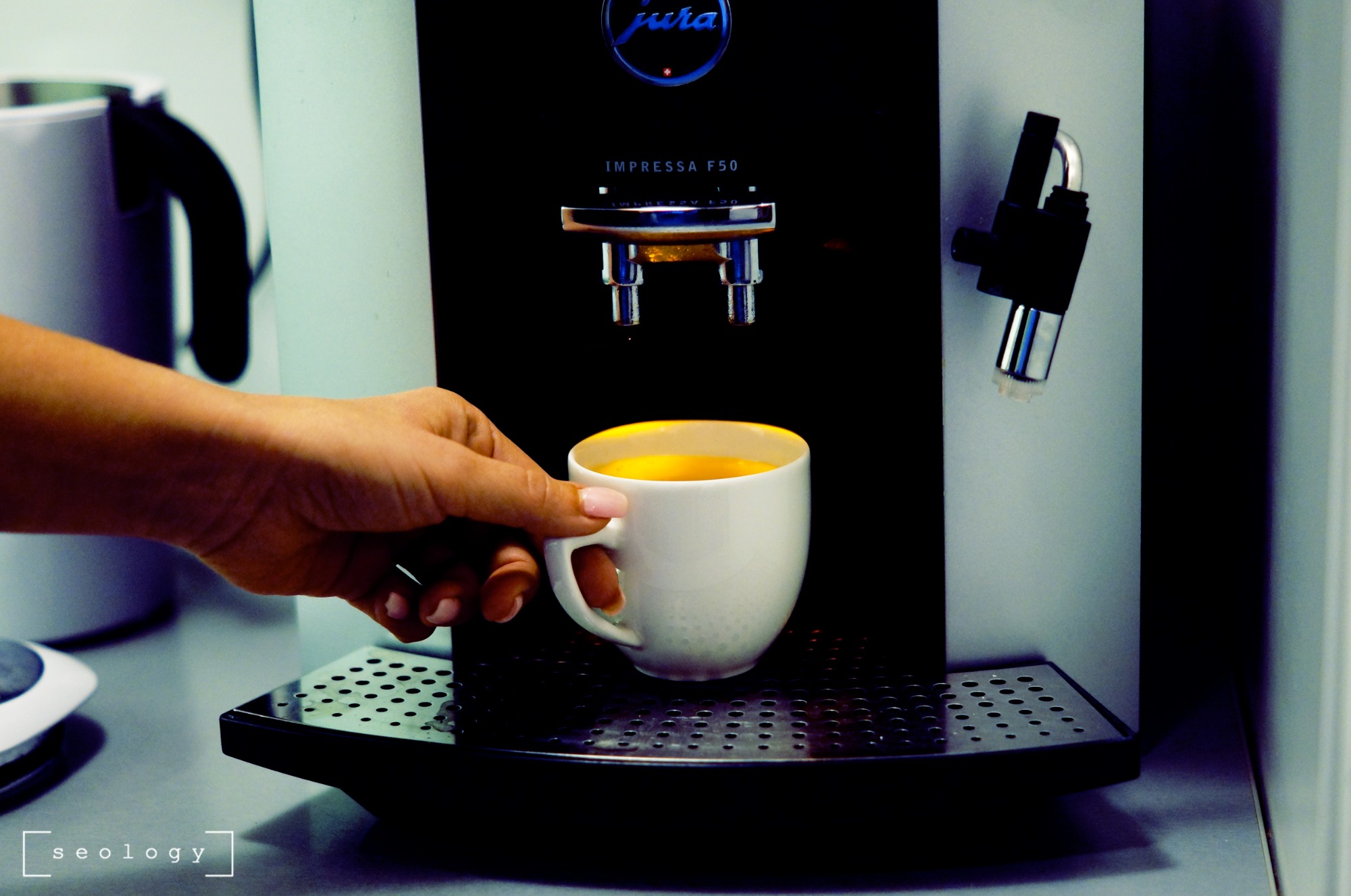 Dłoń sięga po białą filiżankę z kawą z ekspresu Jura Impressa F50 w biurowej kuchni, obok czajnik i blat.