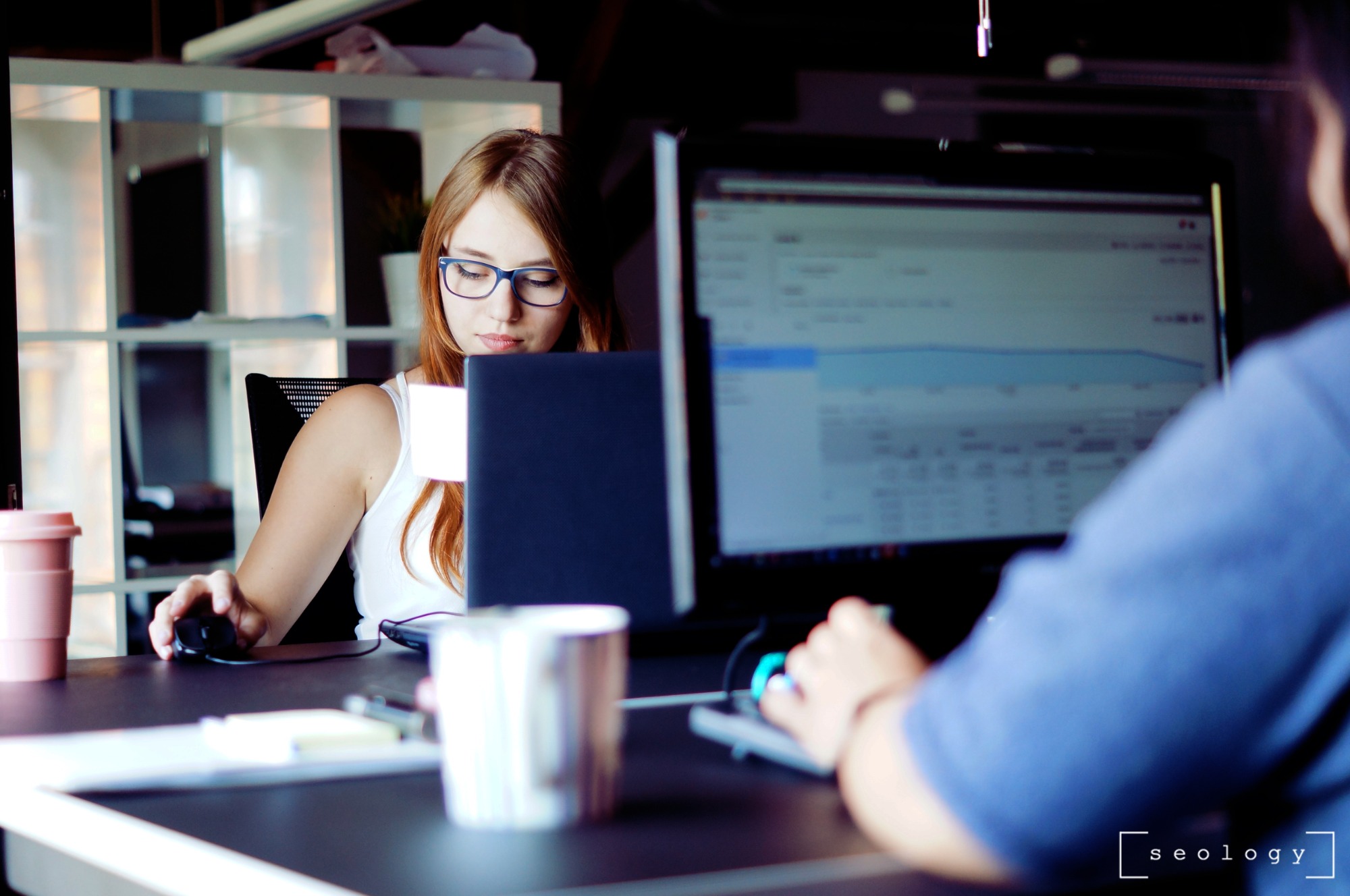 Kobieta w okularach pracuje przy komputerze, na ekranie widoczne wykresy, w tle regał z półkami, w kadrze widoczna również druga osoba. Na biurku kubek z kawą.