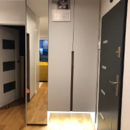 Biała szafa w przedpokoju z lustrzanym frontem, oświetleniem LED u dołu i kalendarzem z kotem na górze, widok na drewnianą podłogę i drzwi wejściowe.